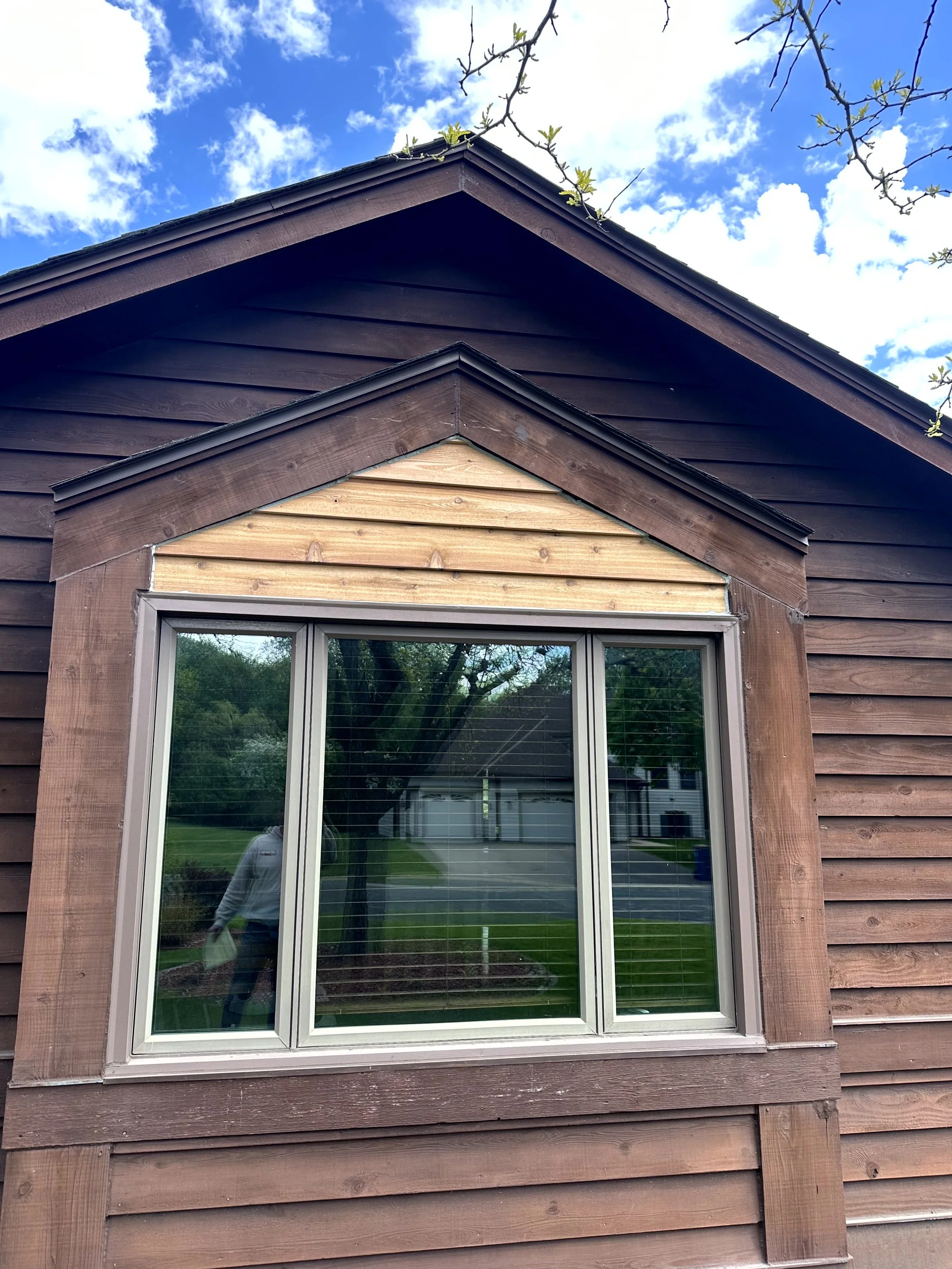 A close-up of a house's exterior showing a window with brown shingles. The window reflects trees and a person taking the photo. The house has a peaked roof with brown and tan siding under the roofline, and a cloudy sky above.