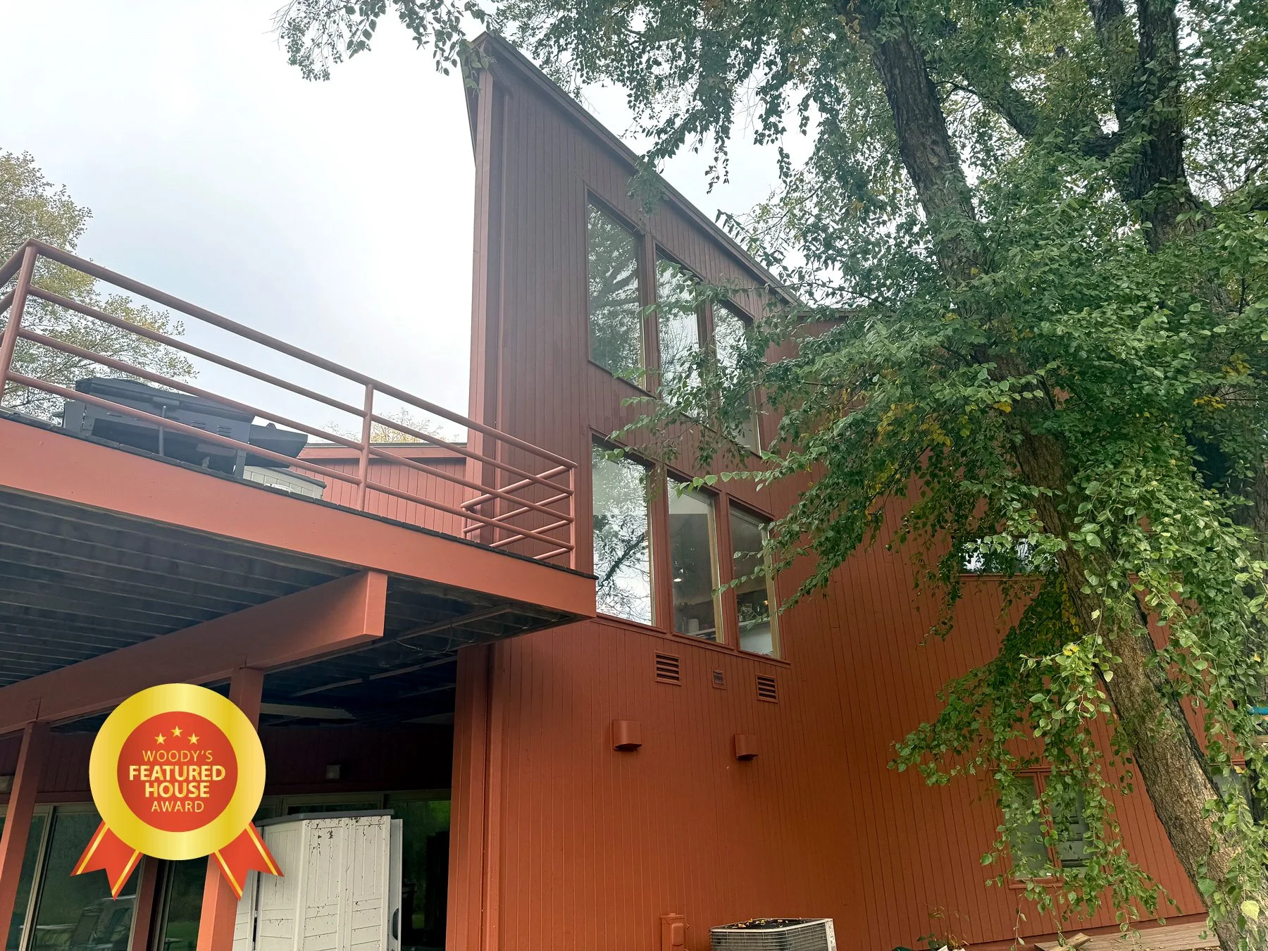 Modern brown multi-story house with large windows and a balcony, surrounded by green trees.