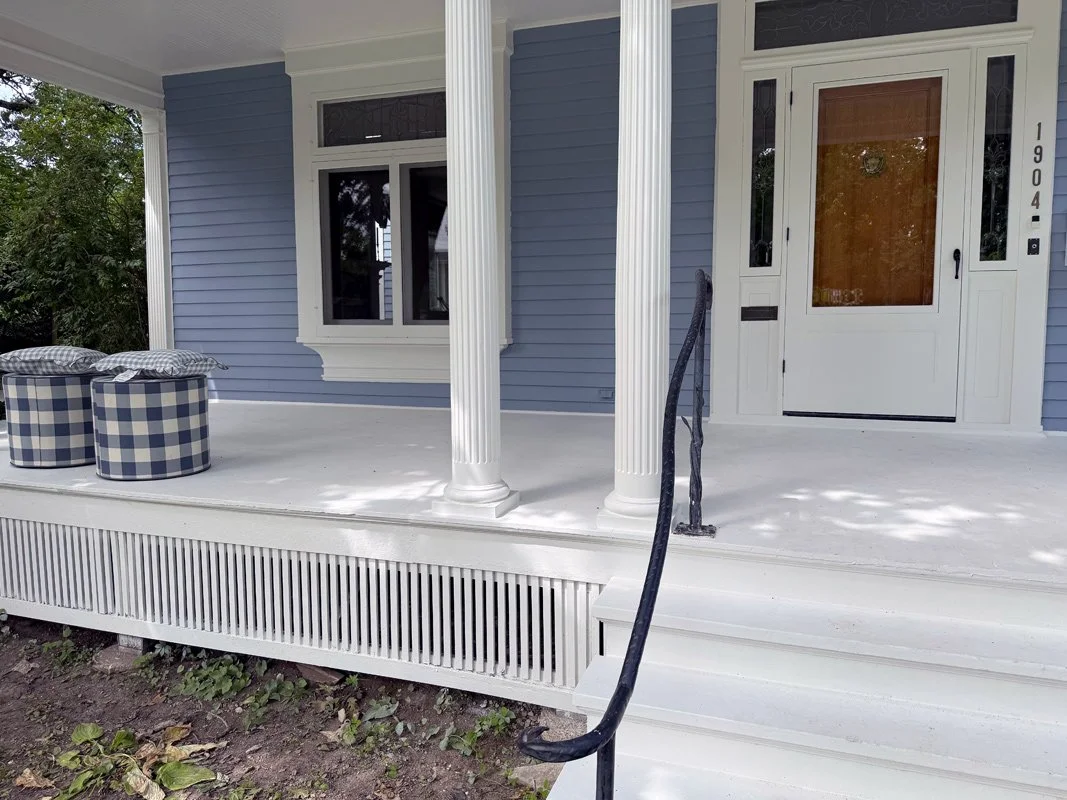 Front porch of a house painted in light blue with white trim, featuring a wooden door with a glass window and house number 1904, two white columns, and outdoor furniture with checkered cushions.