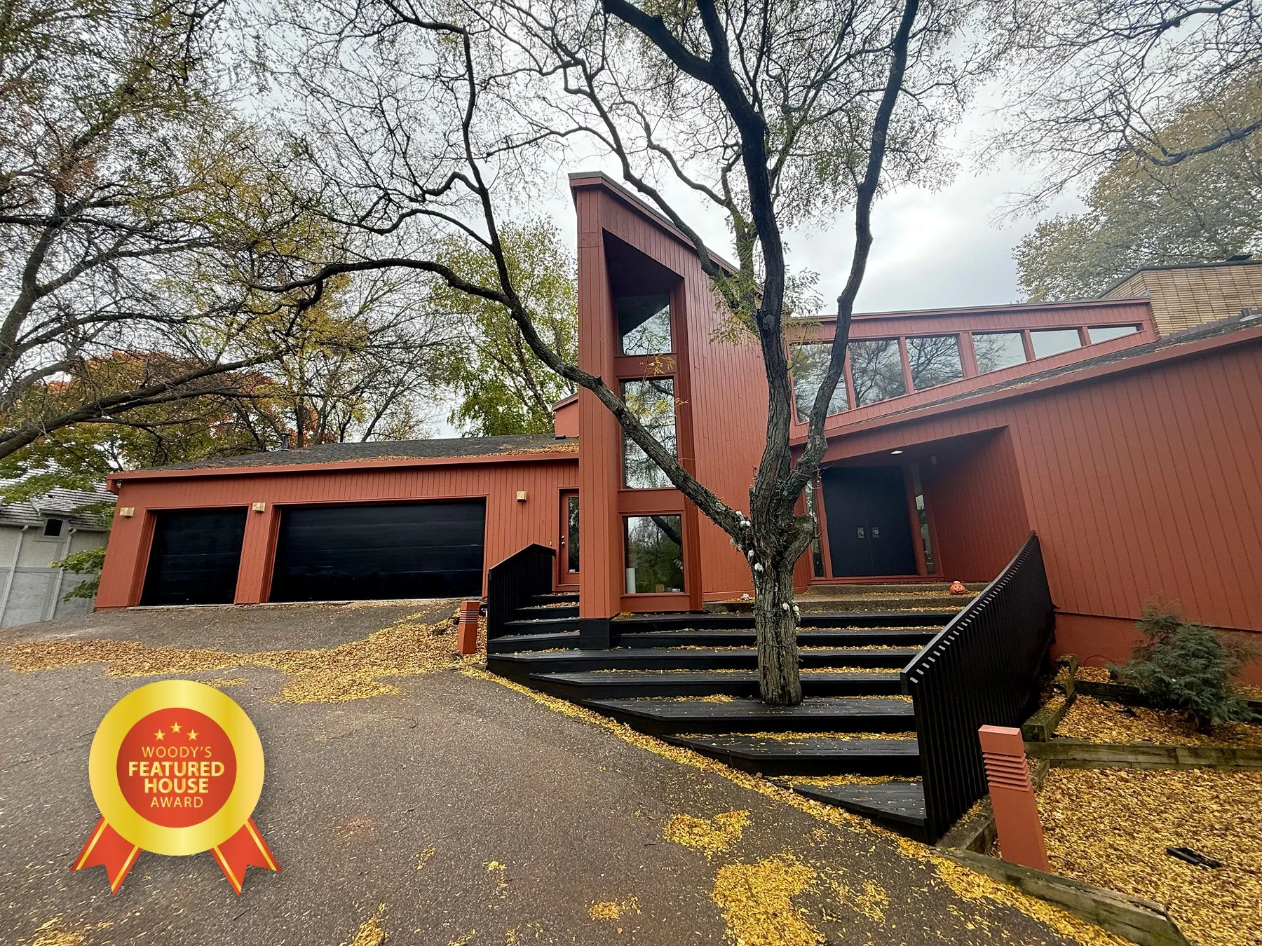 Modern red house with large garage doors, black stairs leading to the front door, and a prominent vertical window in a tree trunk.