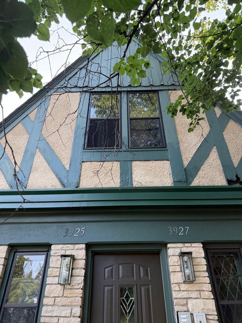 Front view of a two-story house with a stone and stucco exterior, dark front door, decorative glass panel, and a window on the upper level partially obscured by green tree branches.