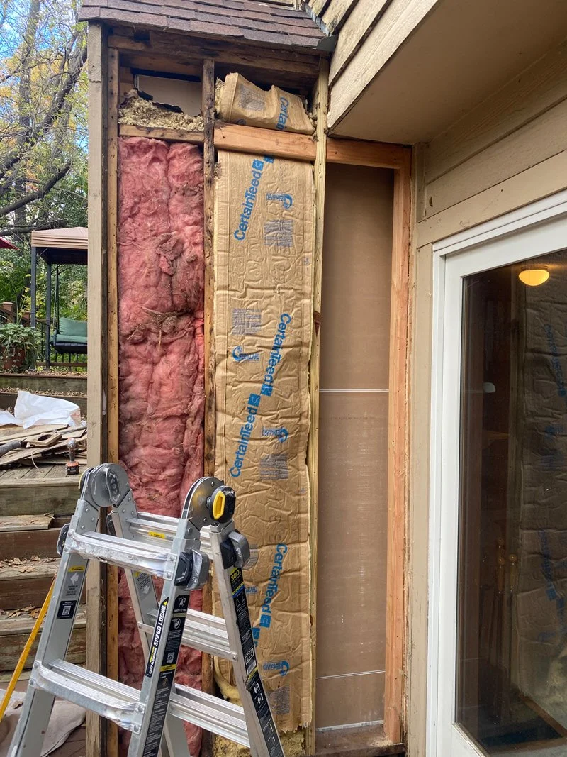 Partial exterior wall of a house under renovation with insulation being installed. Pink insulation and Kraft paper insulation with blue text are visible. A stepladder is in front of the wall, and a sliding glass door is on the right side.