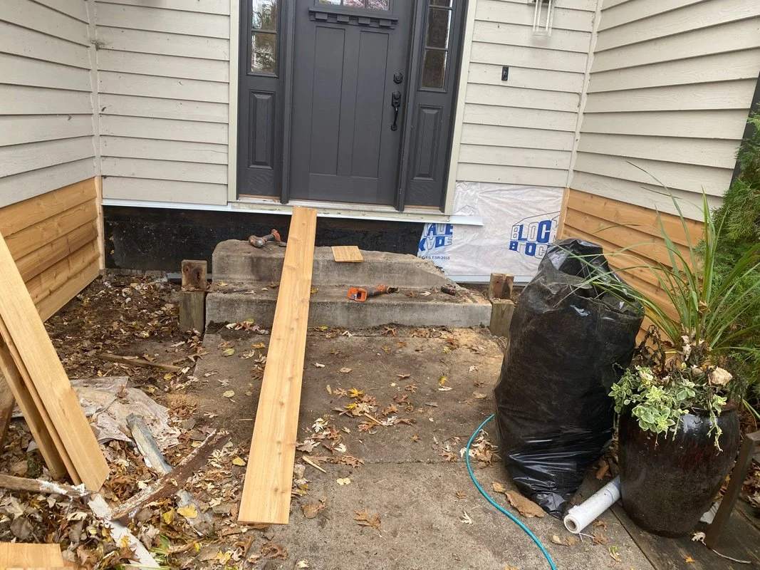 Front porch under construction with wooden boards, construction tools, and supplies, including a black garden pot and a covered bag, in front of a house with beige siding and a black front door.