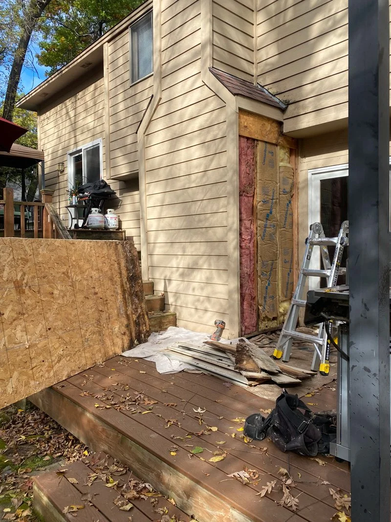 Outdoor construction or repair site with a partially boarded-up door or window, a ladder, construction tools, and plywood sheets scattered on a wooden deck.