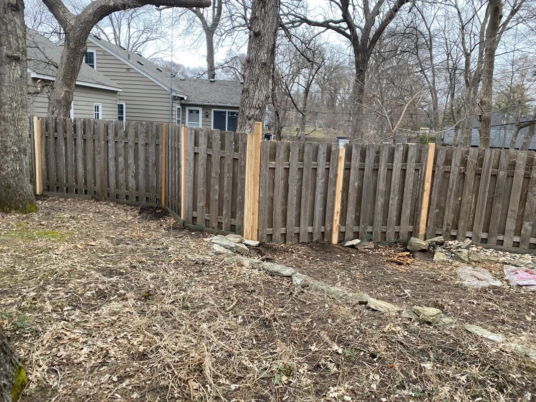 A wooden fence partially repaired with new wood in backyard with trees and a house in the background.