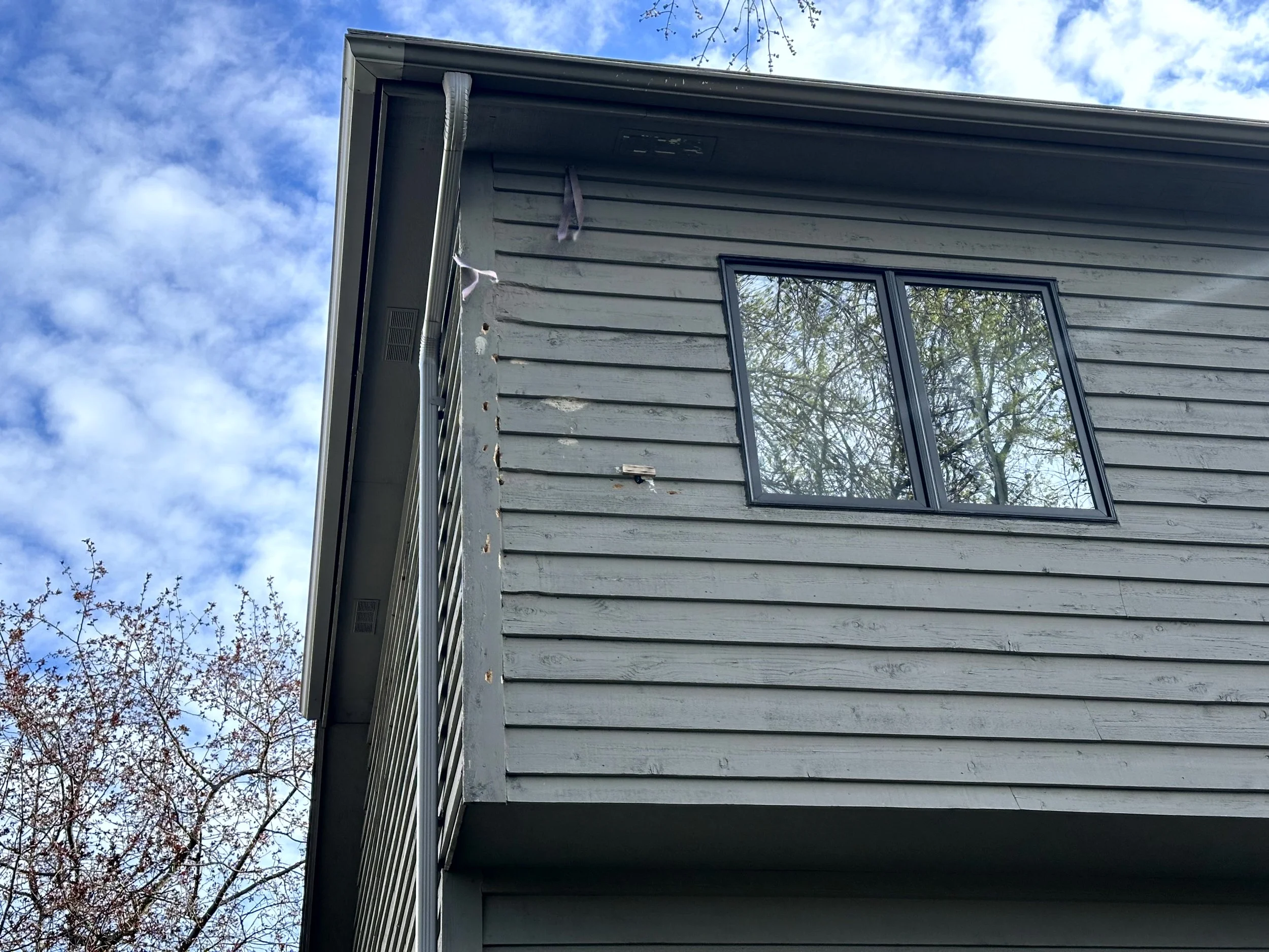 Exterior view of a gray wooden house with trim and a window reflecting trees and blue sky