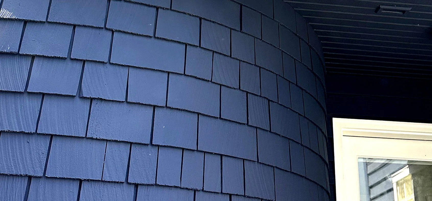 Close-up of dark blue shingle siding on a curved exterior wall near a white-framed window.