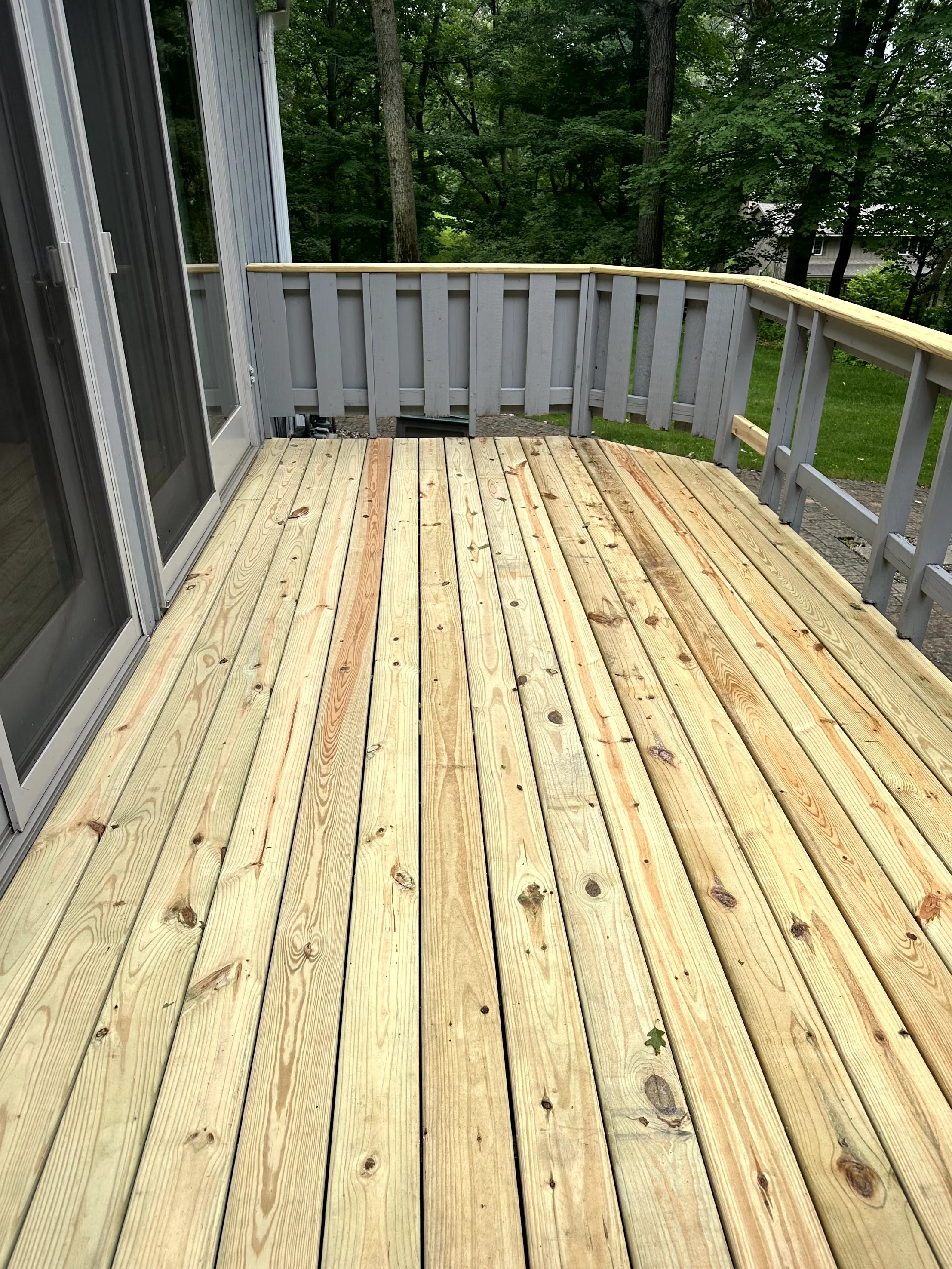 Newly built wooden deck attached to a house, surrounded by a gray painted railing, overlooking a green backyard with trees.