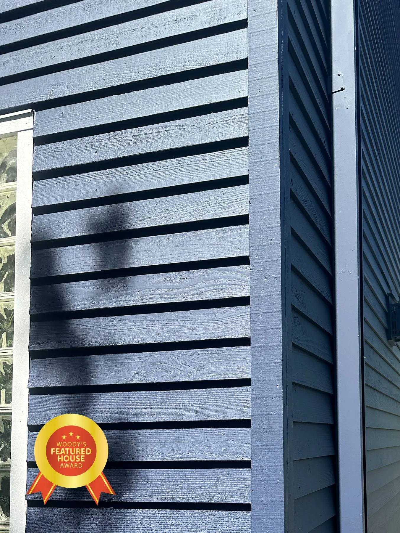 Close-up of a blue house exterior wall with horizontal siding, with a shadow cast on it and a 'Woody's Featured House Award' badge in the bottom left corner.