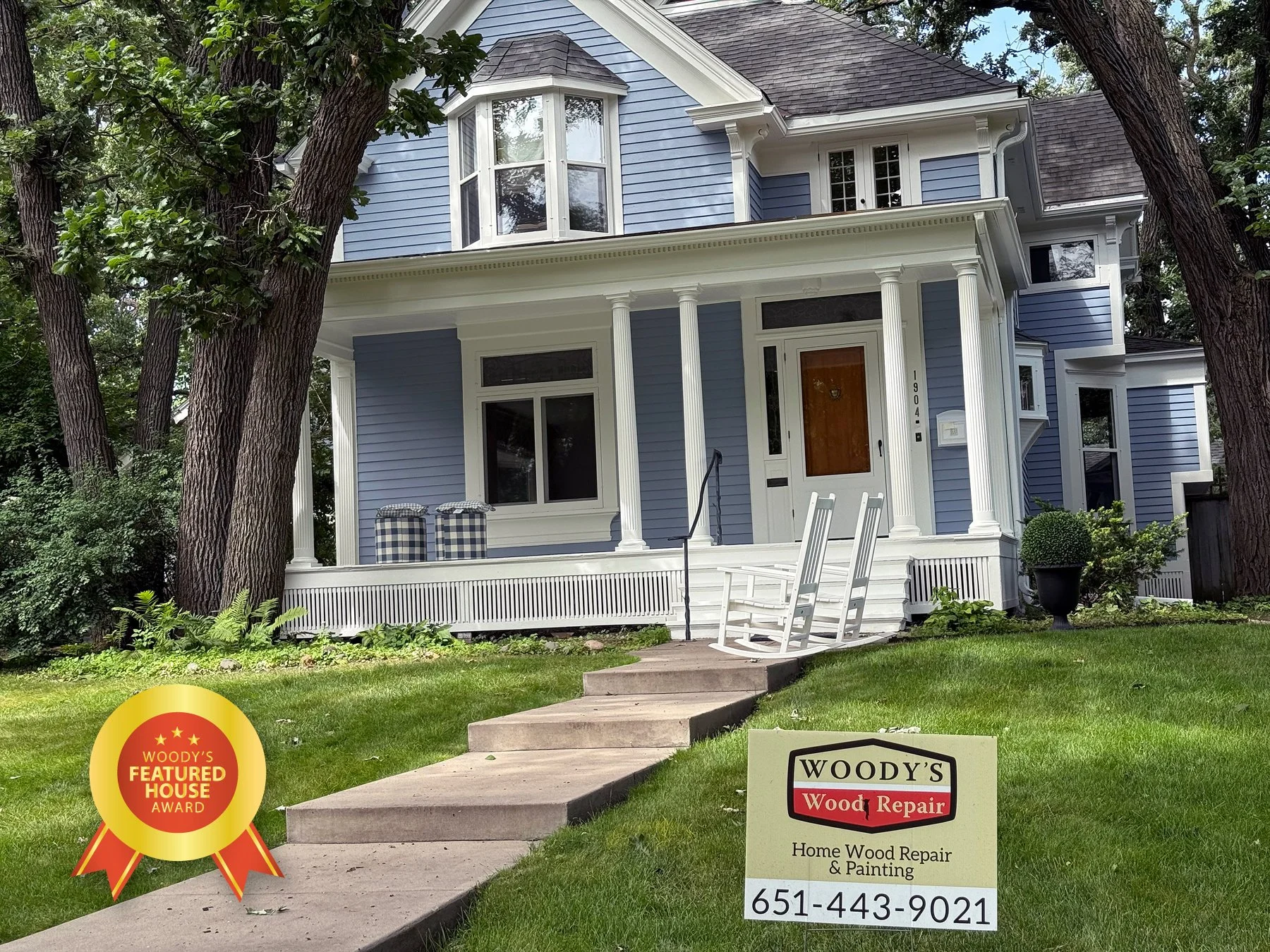 A two-story house painted in light blue with white trim, a front porch with white railing, and large trees in the yard. There is a white rocking chair on the sidewalk leading to the porch. A sign in front of the house advertises Woody's Home Wood Rep