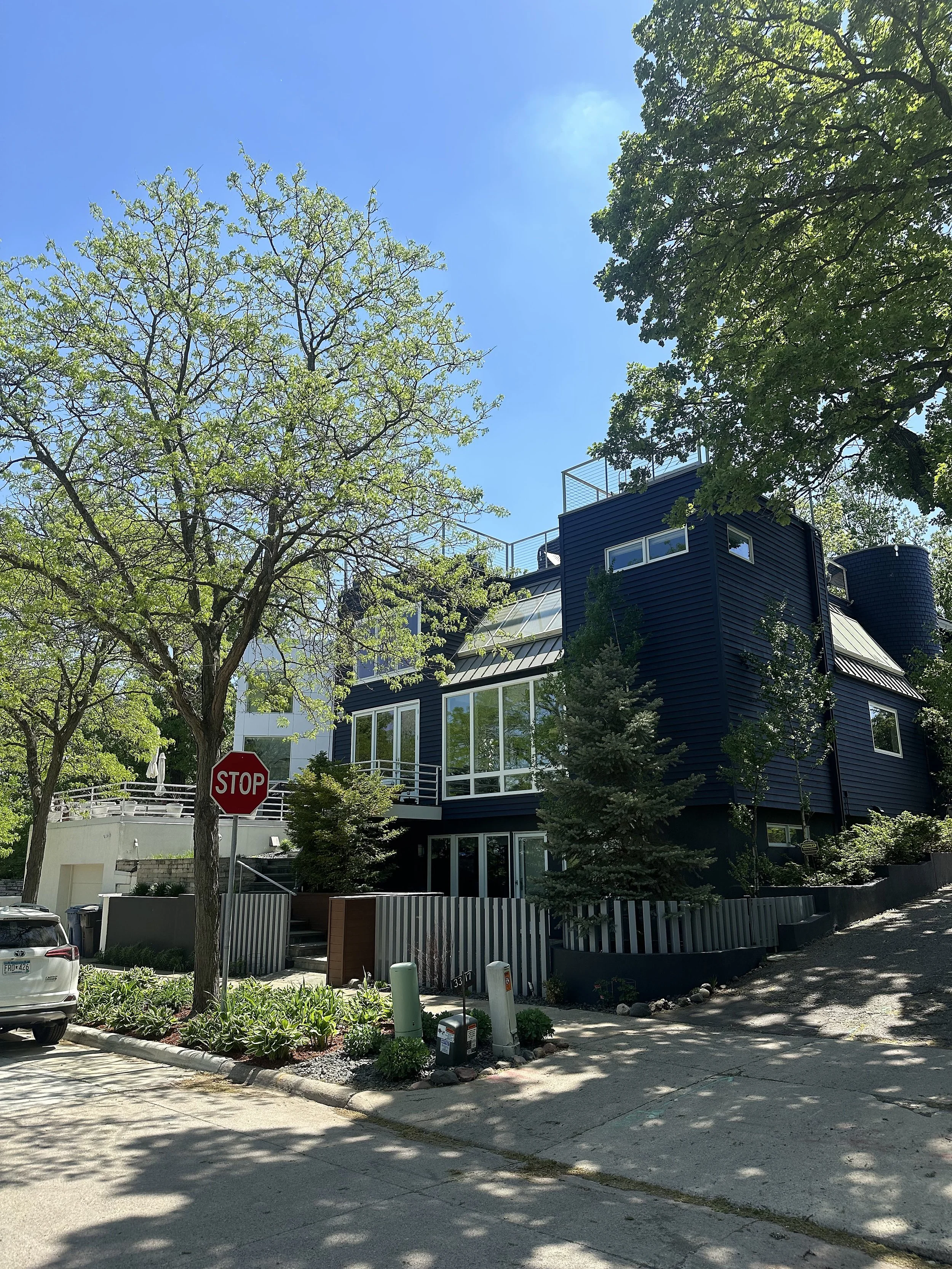 A modern dark blue house with large glass windows in a lush green neighborhood, with trees and a sidewalk in front, and a stop sign nearby on a sunny day.