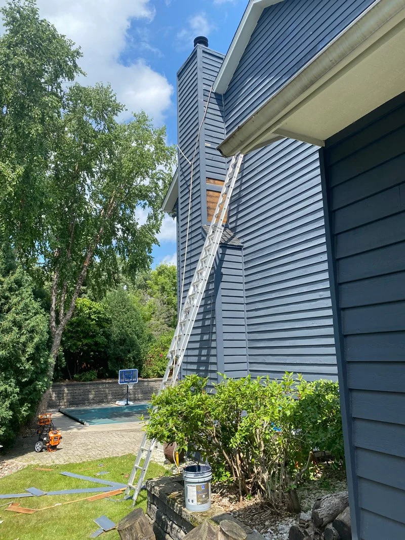 A ladder leaning against the side of a blue house, reaching the chimney. A shrub, a bucket, and some tools are in the yard. There is a basketball hoop and a trampoline in the background.