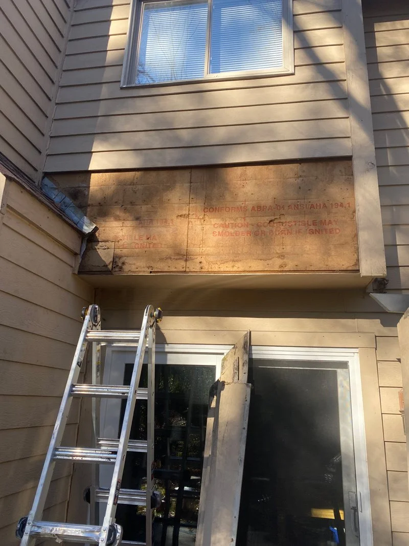 Exterior view of a house under renovation with a ladder leaning against the wall, a piece of plywood covering part of the wall with red warning text, and a window above.