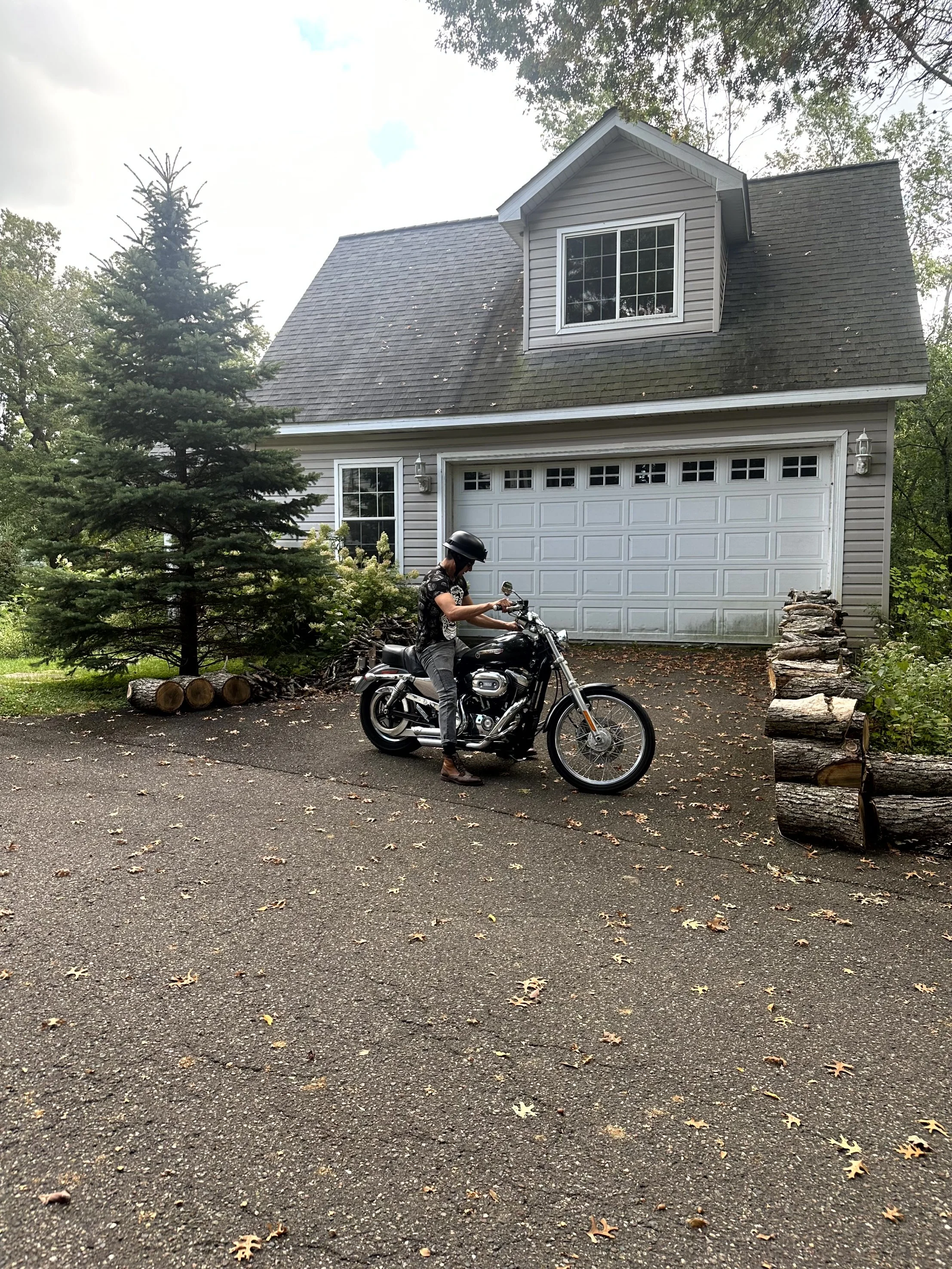 A woman wearing a helmet sitting on a black motorcycle in front of a two-story house with a garage, surrounded by trees and fallen leaves.