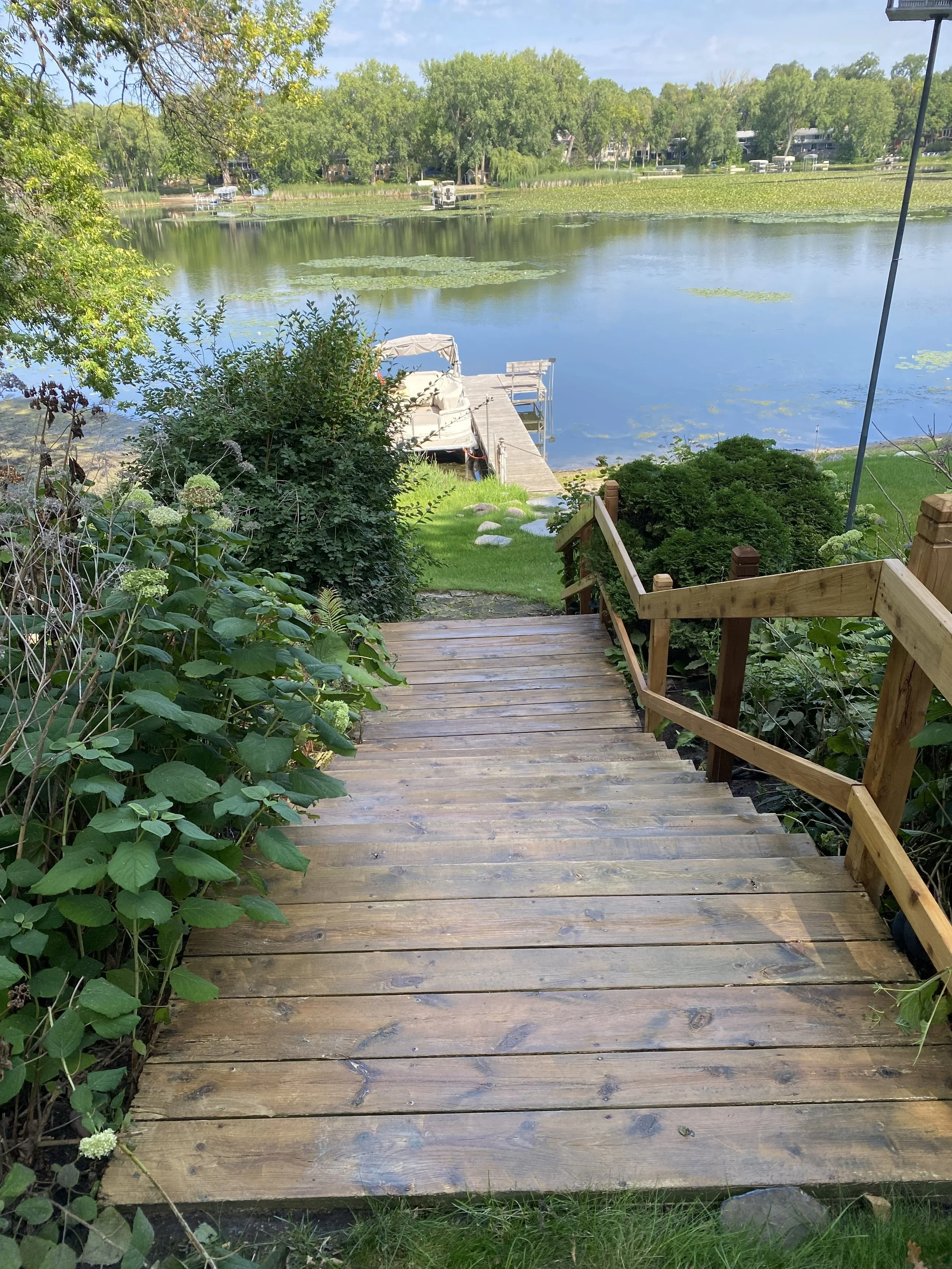 Wooden staircase leading down to a dock on a lake, with a boat attached, surrounded by greenery and trees, and a calm water surface with lily pads.