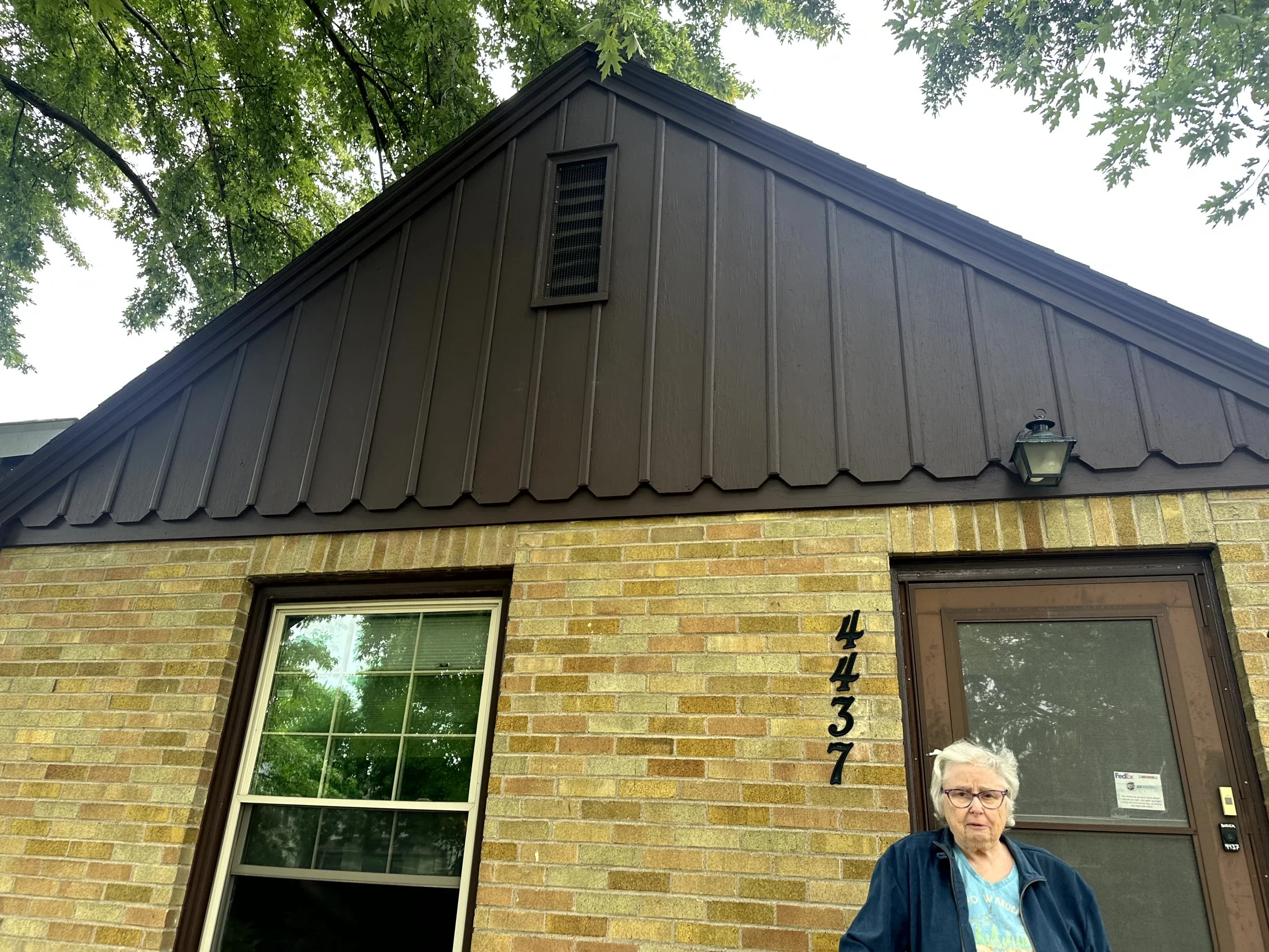 A brick house with a black metal roof, a window reflecting green trees, and a woman with glasses standing outside near the front door.