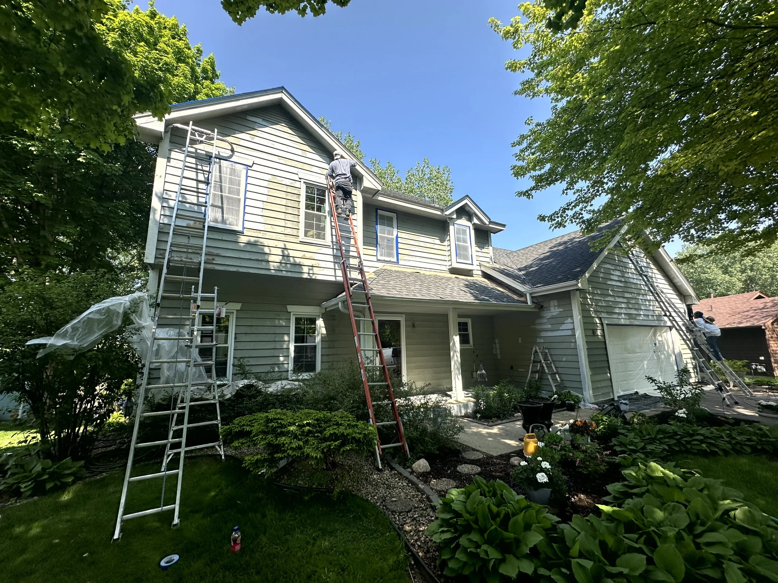 House being painted or repaired with workers on ladders, surrounded by lush green trees and garden plants, under a clear blue sky.