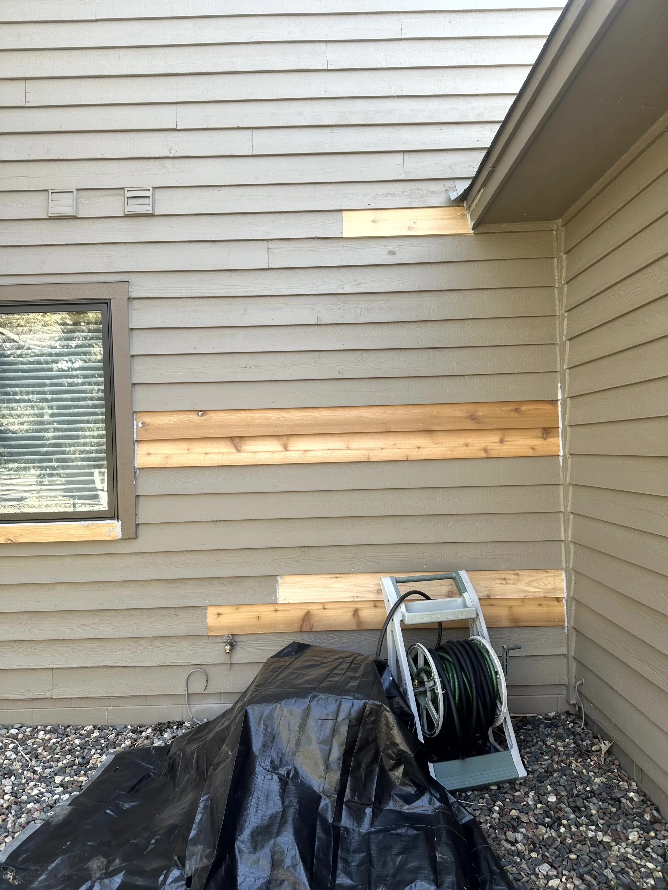 Exterior view of a house wall under construction with new wooden siding boards, a window with blinds, and a garden hose reel on the ground covered partially with a black tarp.