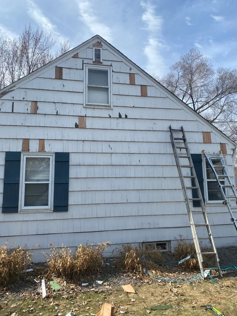 A house with peeling white siding and two windows with blue shutters; ladder and repair tools leaning against it, and dried plants in front.