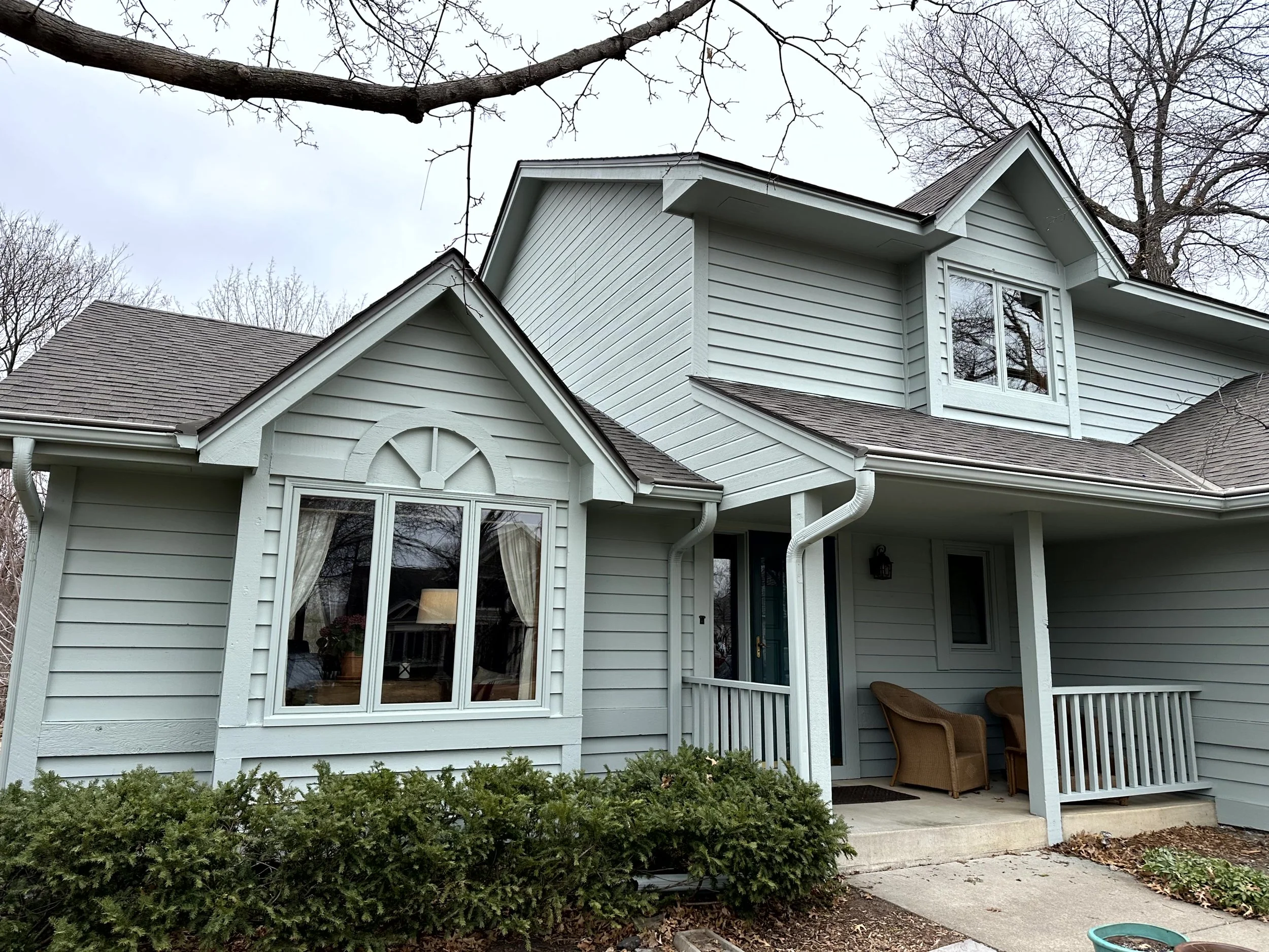 A two-story house with light blue siding, white trim, and a gabled roof, featuring a front porch with two chairs and a small railing, and a large front window with curtains, surrounded by leafless trees and bushes.
