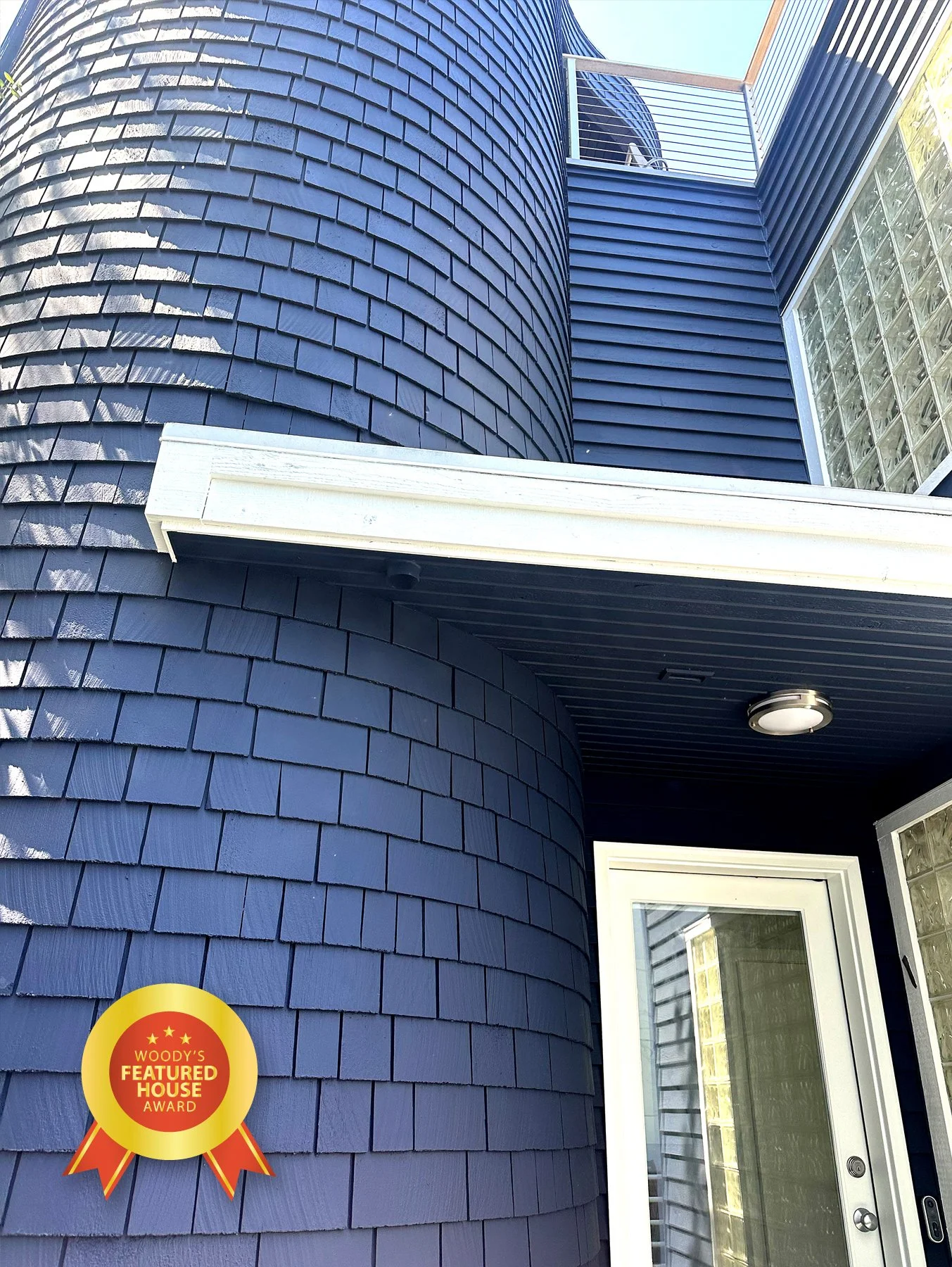 Close-up view of a blue house exterior with shingle siding, a glass block window, a sliding glass door, and a balcony with railing. An award badge reading 'Woody's Featured House Award' is in the lower left corner.