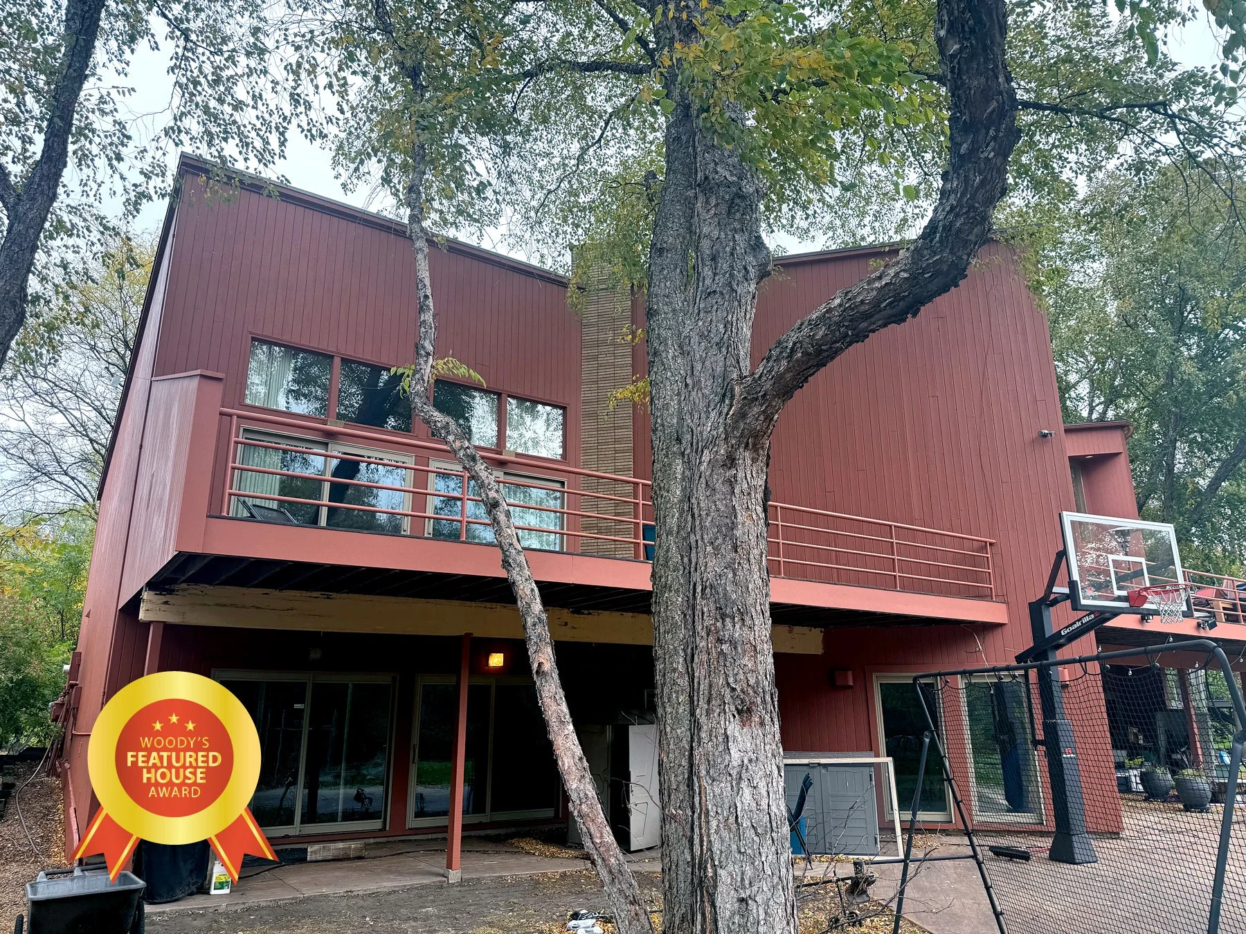 Modern multi-story house with red vertical siding, large windows, and a balcony with metal railing, surrounded by trees, with a basketball hoop and outdoor equipment in the backyard.