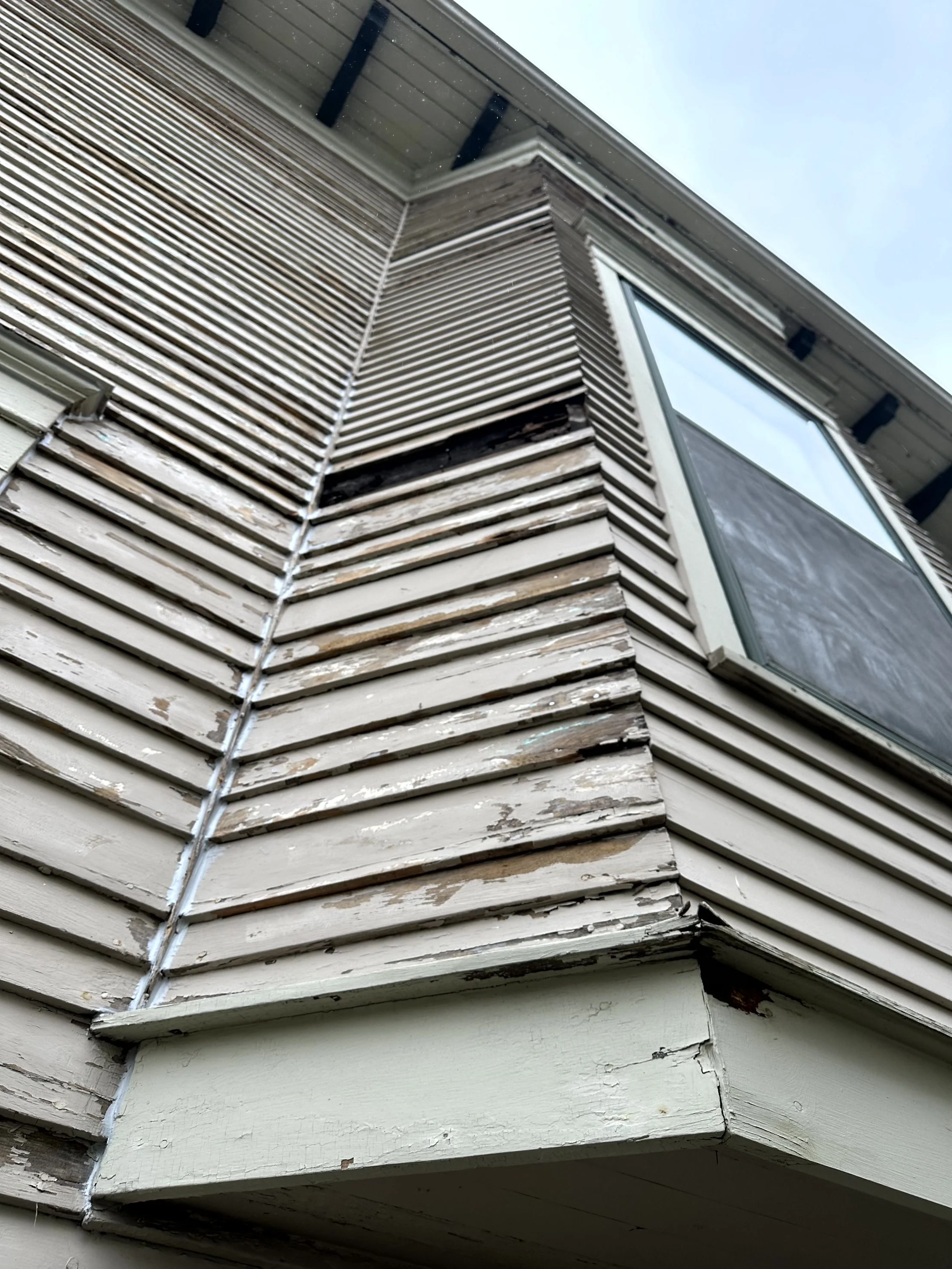 Close-up of a corner of a house with peeling beige siding, showing signs of weather damage and wood rot near the bottom edge.