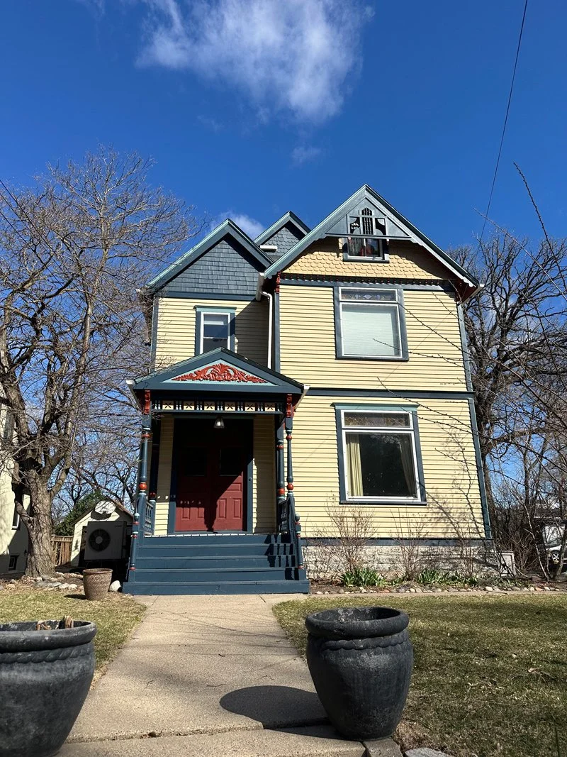 A three-story Victorian house with yellow siding, blue accents, and ornate trim. The house has a red front door, a small porch with steps, and large windows. The sky is clear with a few clouds, and leafless trees surround the house.