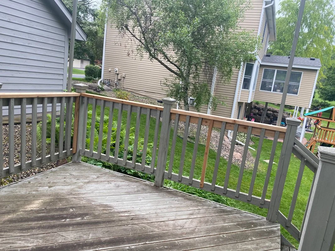 View of a backyard deck with gray railing, adjacent to a garden and neighboring house with beige siding, trees, and a playset with a slide in the background.