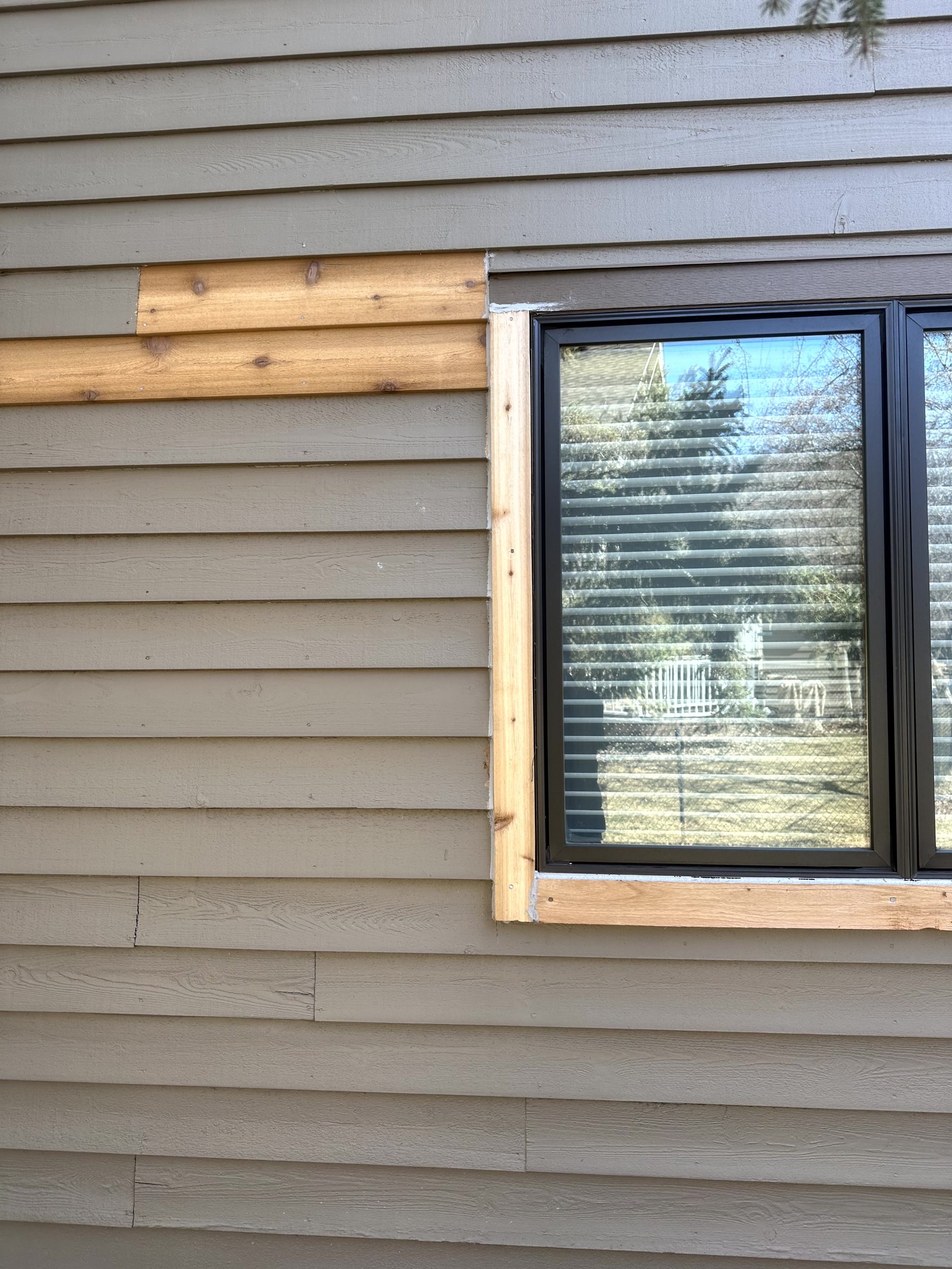 Exterior wall of a house under construction with horizontal siding, partially completed window frame with black window, and some wood trim around the window.