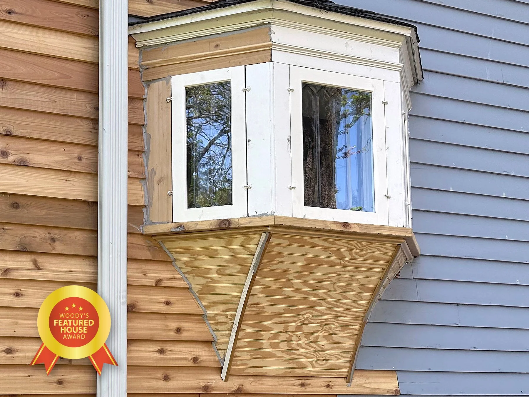 Newly constructed bay window with white frame on a house with two-tone siding, featuring a wood underside and a metallic gutter.