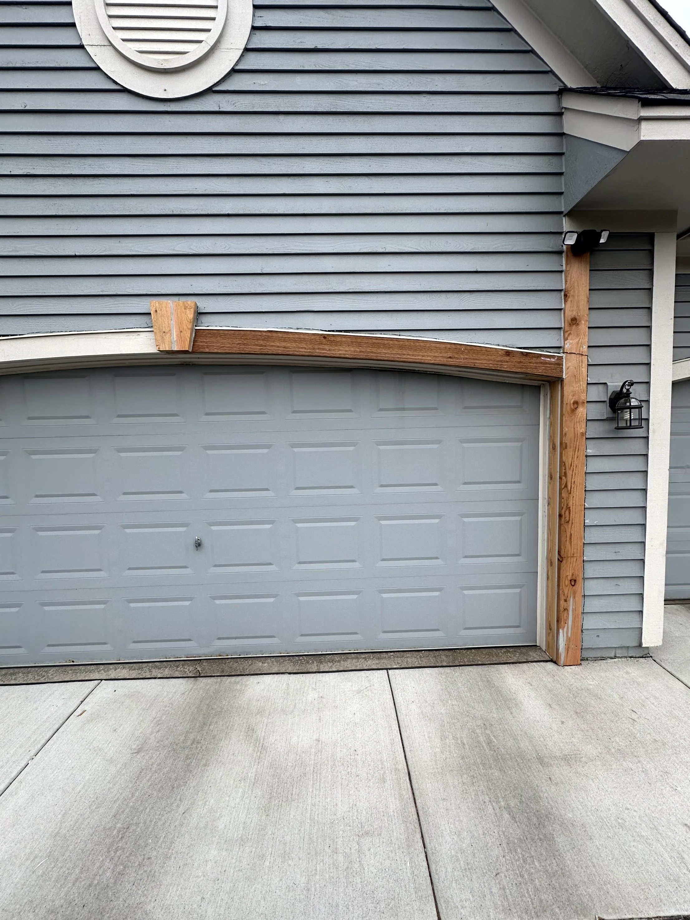 The front of a residential house with a closed garage door, blue siding, and decorative details. There is a new wooden post on the right side and a wooden piece on the top of the garage door, possibly for future repair or decoration. A small black ou