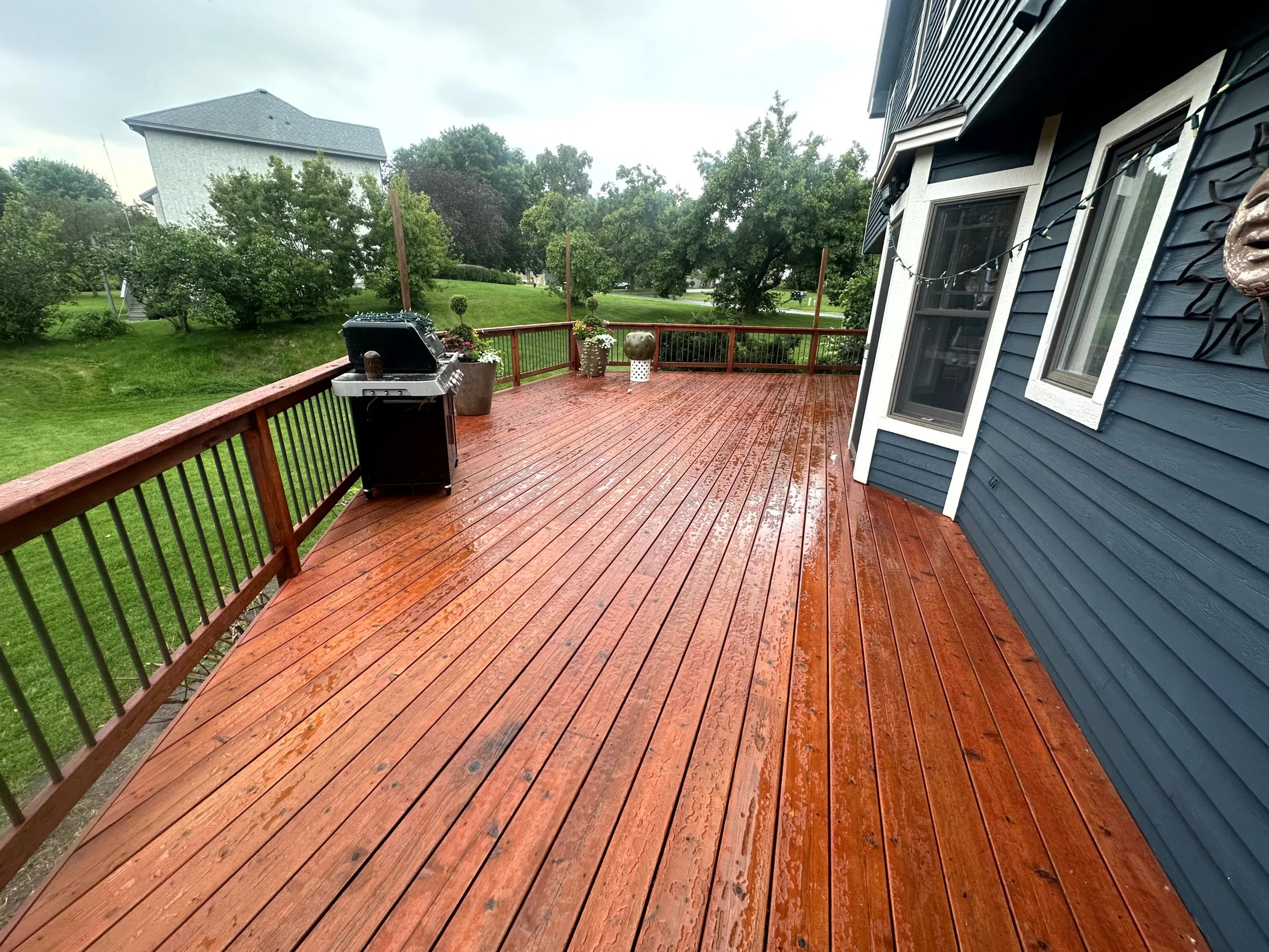 Wet wooden deck with a grill, potted plants, and house exterior with blue siding on a rainy day, overlooking a grassy yard with trees.