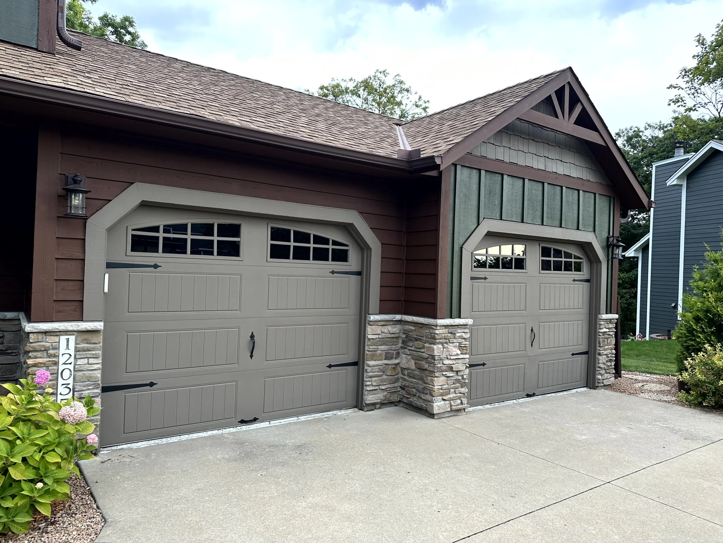Two-car garage with tan doors, stone accents, and a red and green exterior, situated in a residential neighborhood with landscaping and neighboring houses.