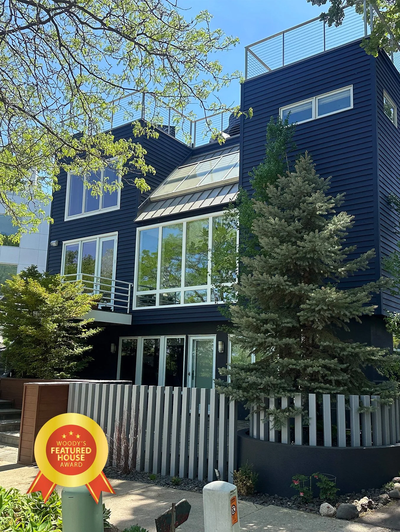 A modern multi-story house with dark blue siding, large glass windows, and a small balcony. There is a tall pine tree in front, a wooden and metal fence, and a street sign with a 'Woody's Featured House Award' badge.