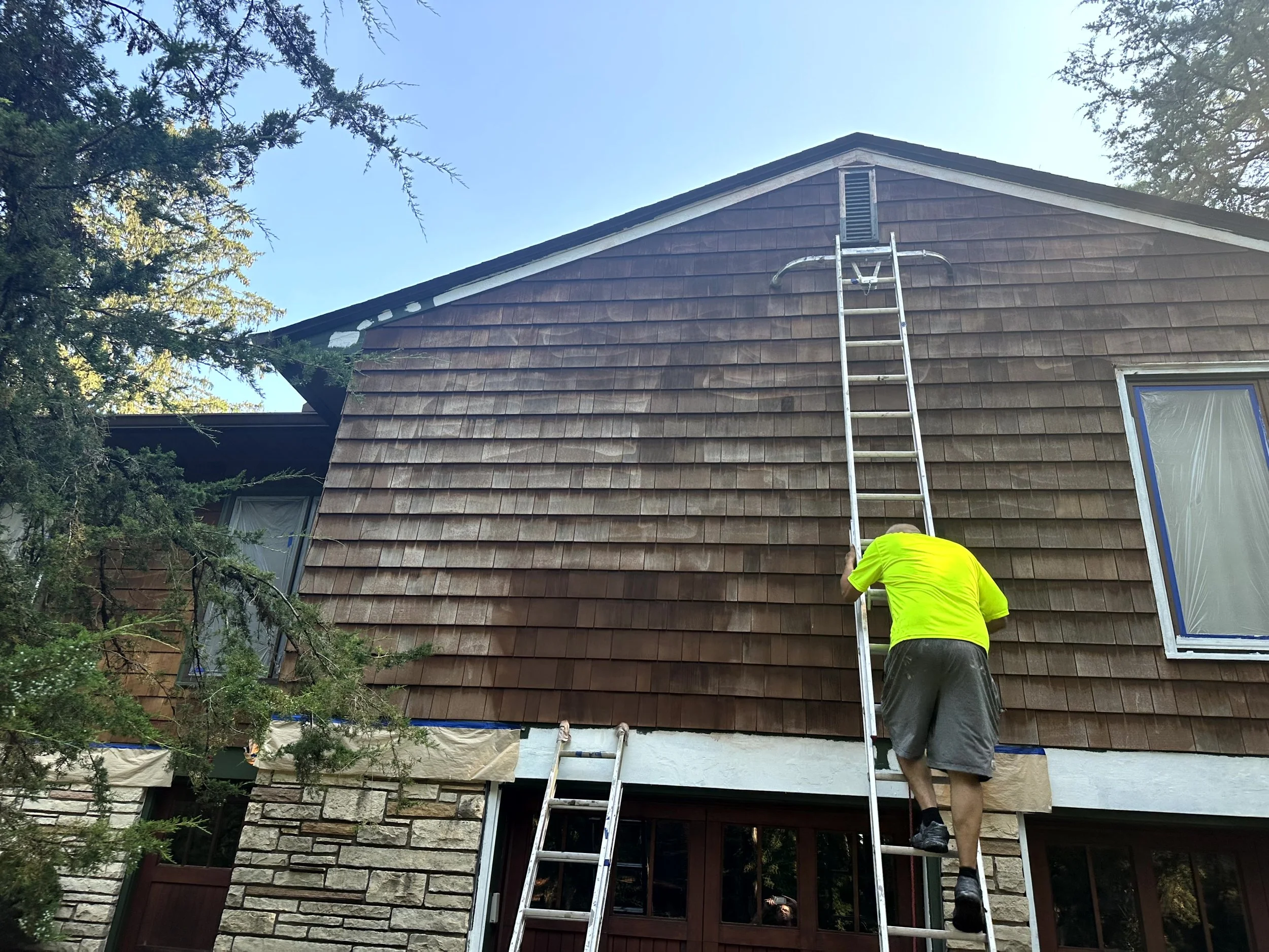 Person in yellow shirt and shorts installing brown wooden shingles on the exterior upper wall of a house using a tall ladder.