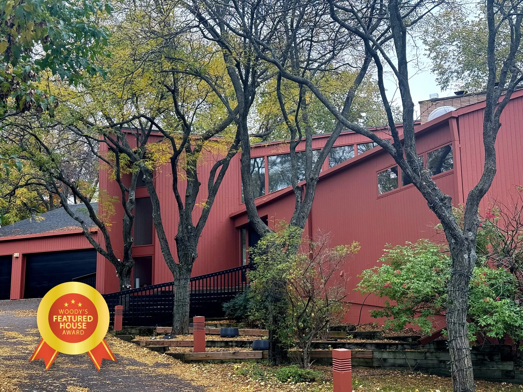 A modern red house with large windows surrounded by trees with yellow and green leaves, indicating fall, and a black railing along the driveway.