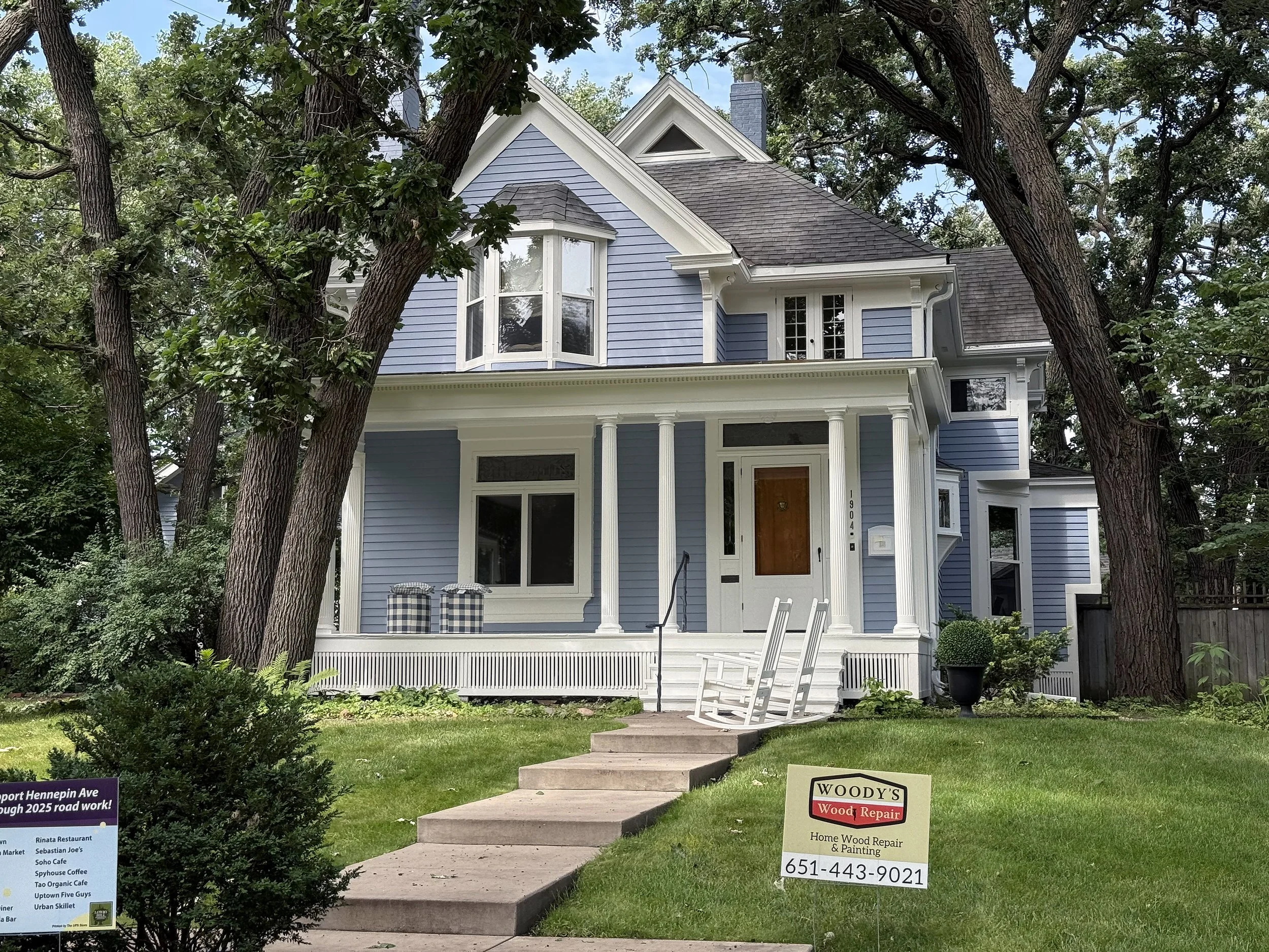 A two-story blue house with white trim, a porch with white columns, a small set of stairs, a rocking chair, and trees surrounding the yard. A wood repair sign and a neighborhood sign are in the grass.