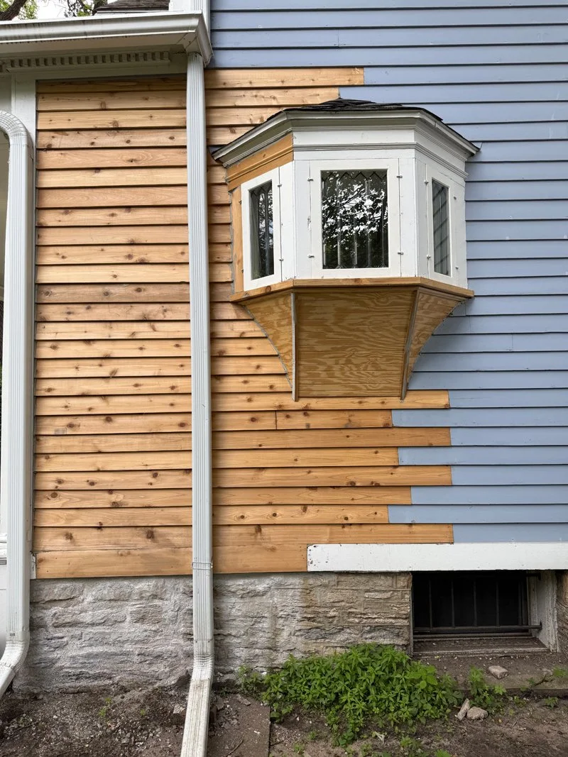 Partial view of a house exterior with blue siding and natural wood paneling, featuring a decorative bay window with white trim, and a concrete foundation with a small basement window. Green plants grow at the base.