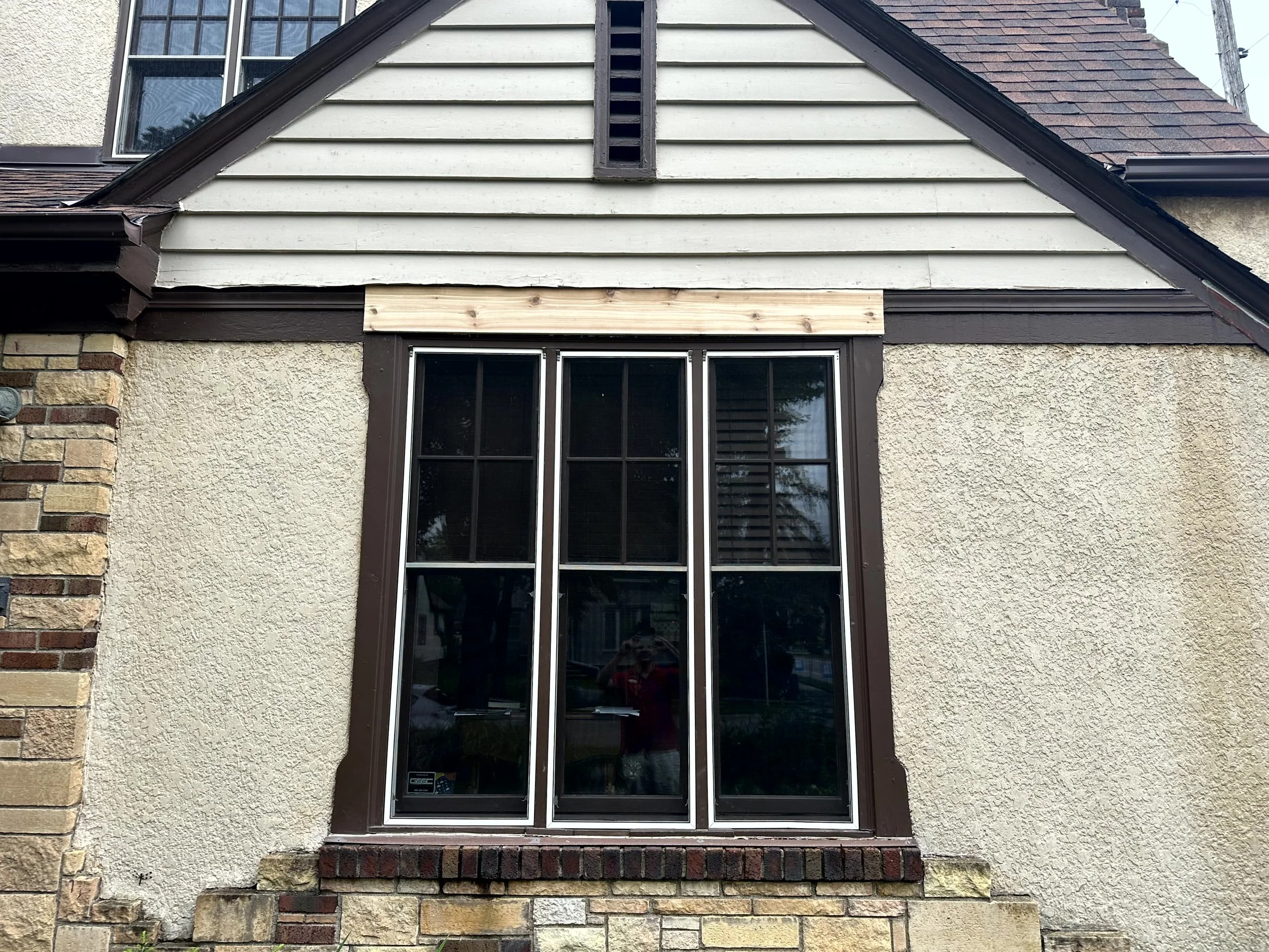 Exterior of a house showing a window with brown trim, part of a beige stucco wall, brick veneer, and a section of siding under a gable roof.