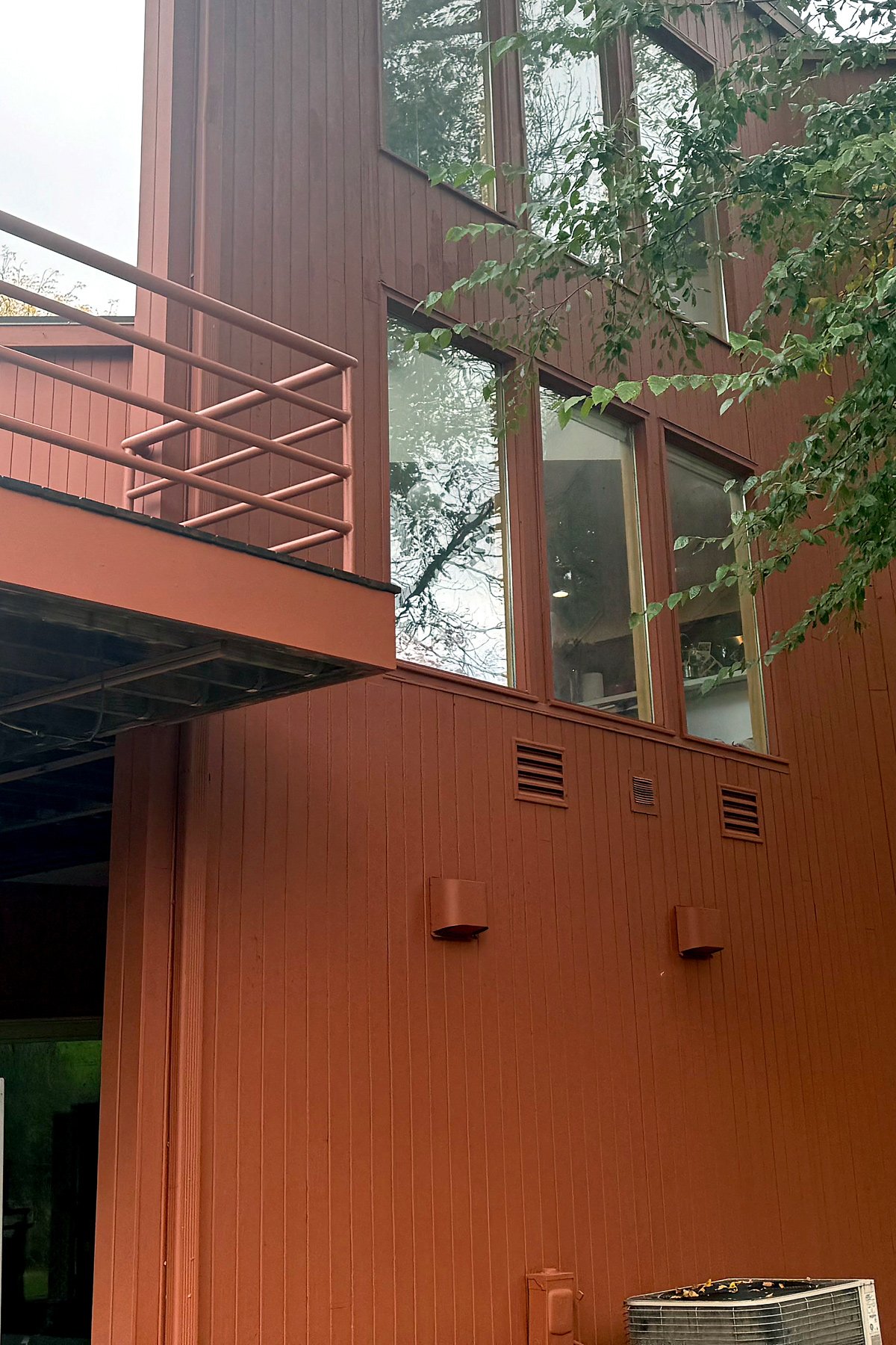 A close-up exterior view of a modern building with a rust-colored, wood-paneled facade, large windows, and a pink metal balcony rail, surrounded by green leafy trees.