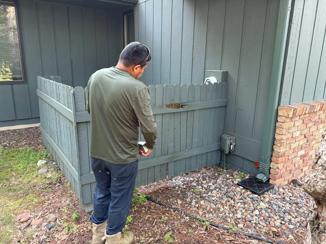 A man with sunglasses and a green jacket standing outdoors near a blue wooden fence and house, holding a tool or device, looking down at the ground.