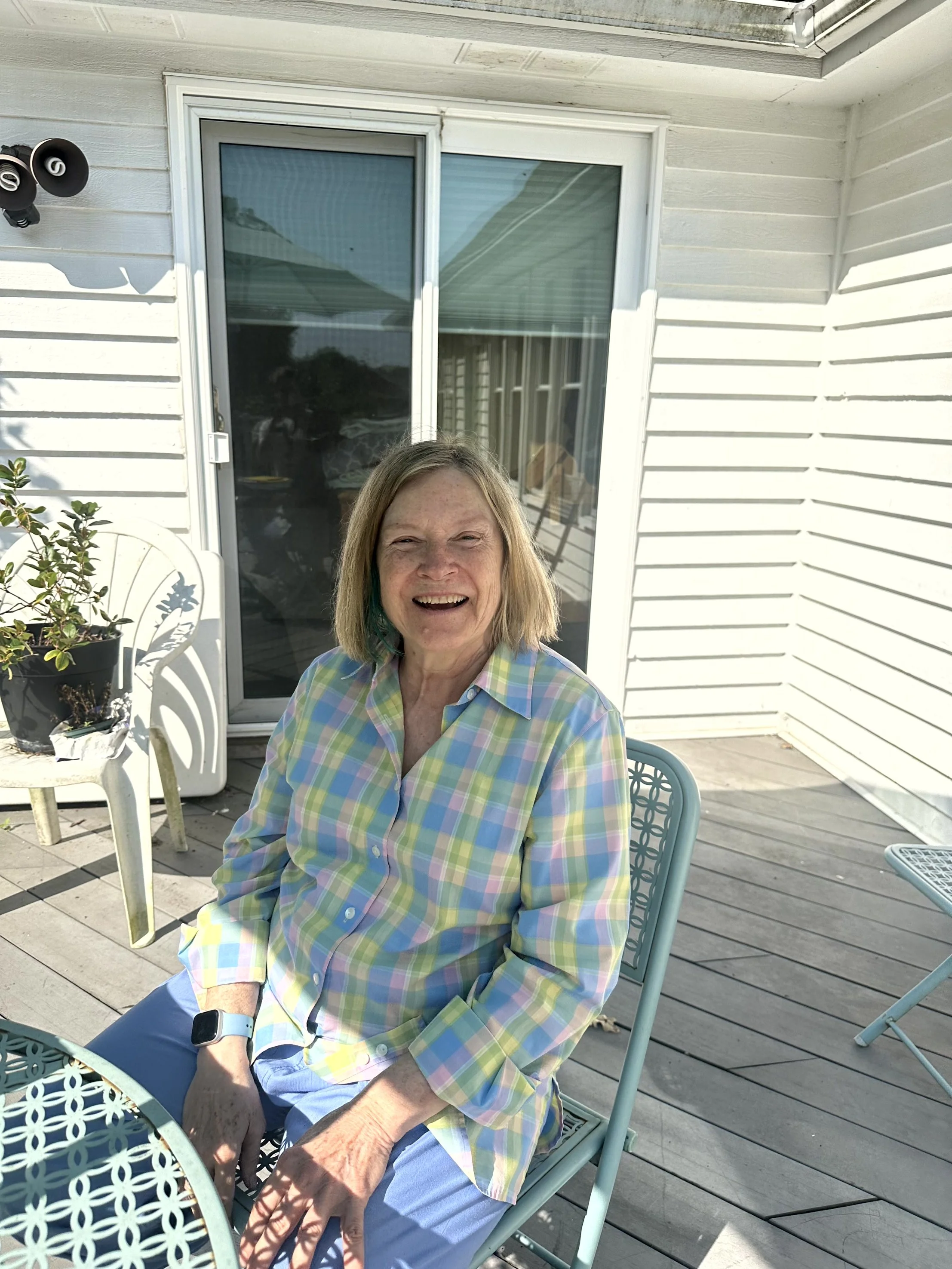 A woman in a pastel plaid shirt sitting outside on a patio, smiling, with a white house and sliding door in the background.