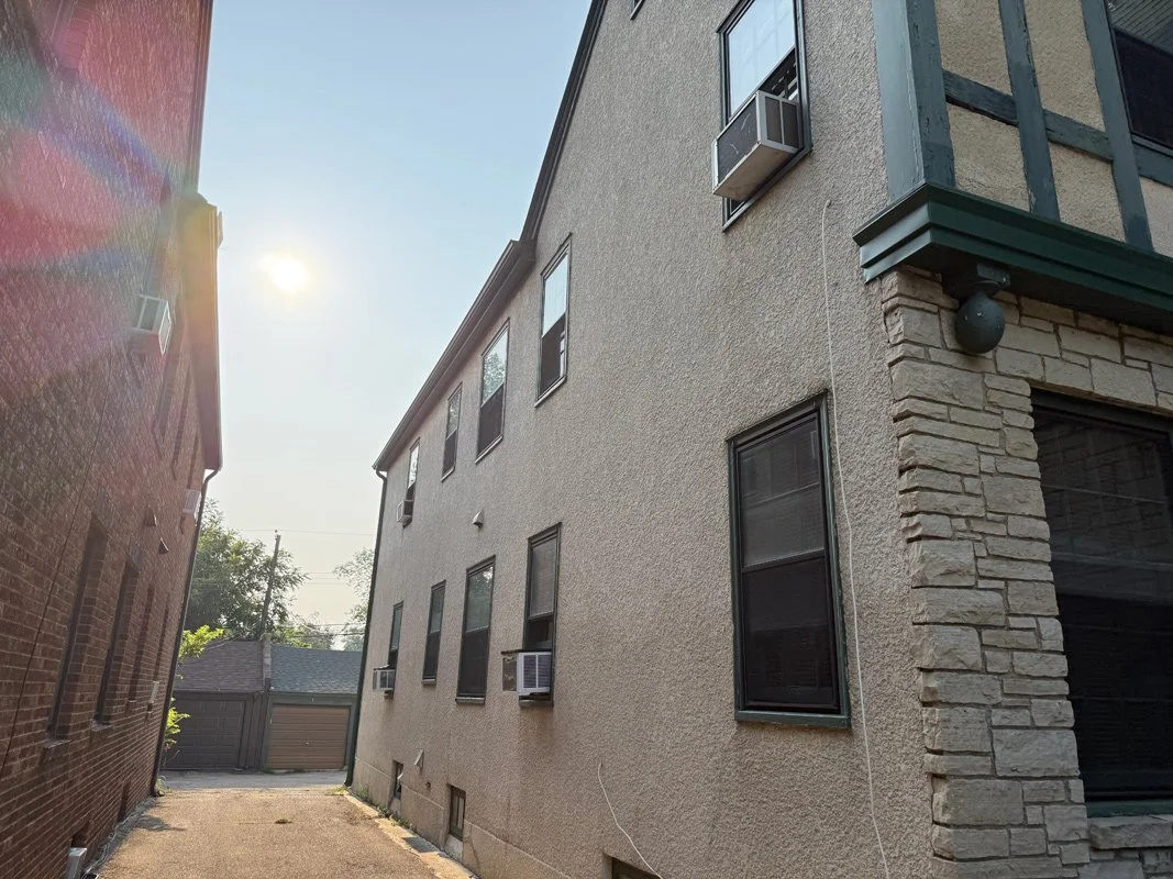 Side alley between a brick building and a beige stucco building with several windows, some with air conditioning units, under a sunny sky. The stucco building was recently painted. 