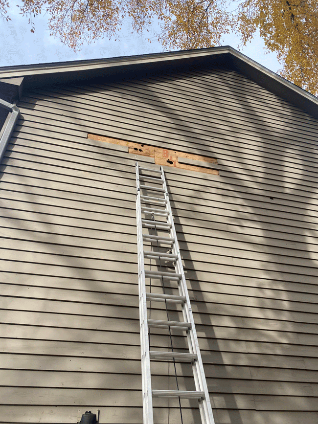 A tall aluminum ladder leaning against the beige horizontal siding of a house, reaching a wooden board that appears to be a missing window or vent opening in the house's upper wall under the roof. The sky and leaves of a tree are visible at the top o