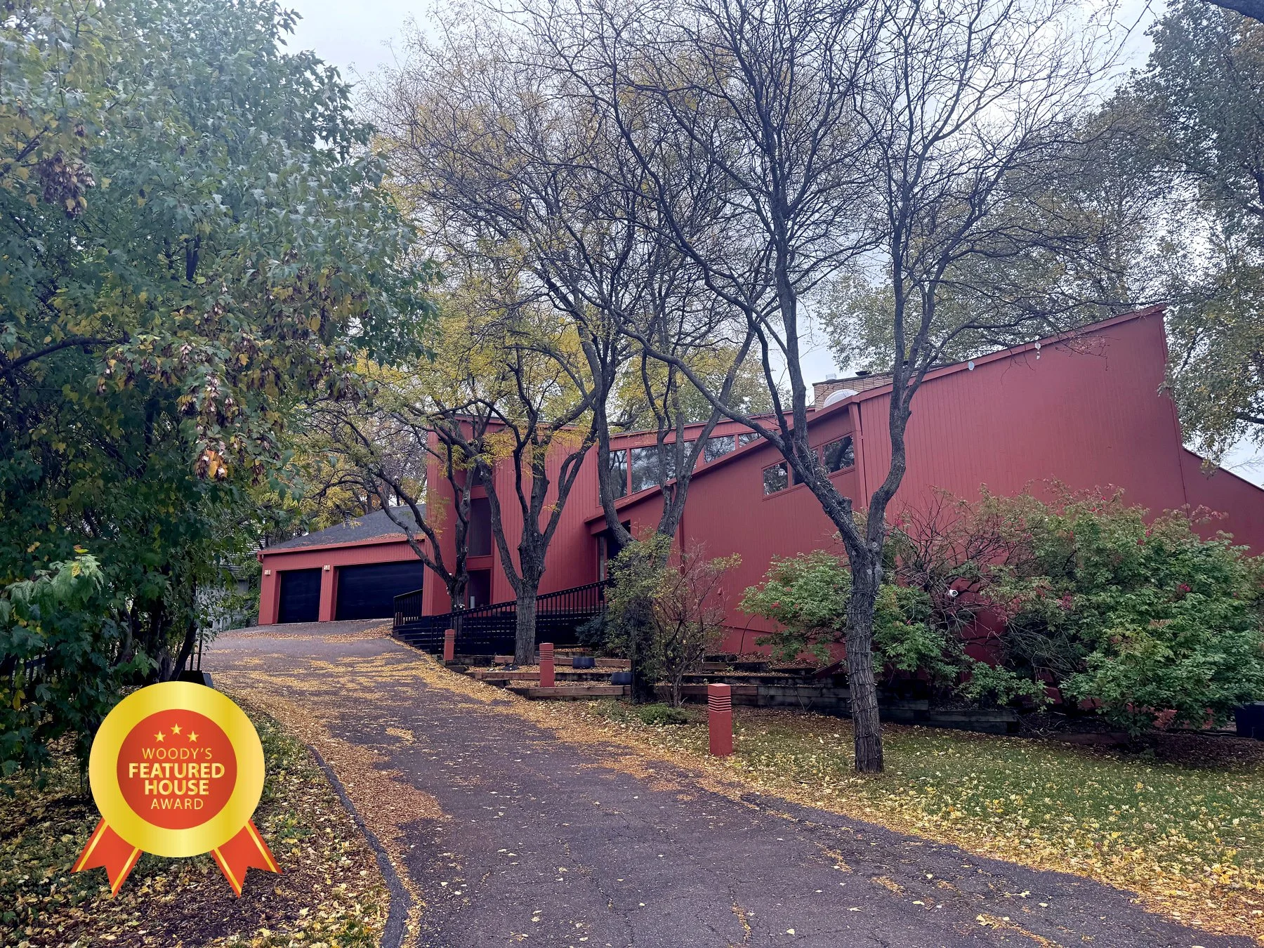 A modern house painted in red with a sloped roof, two garage doors, and large windows, surrounded by trees with autumn foliage and a driveway with fallen leaves, featuring an award badge at the bottom left corner.