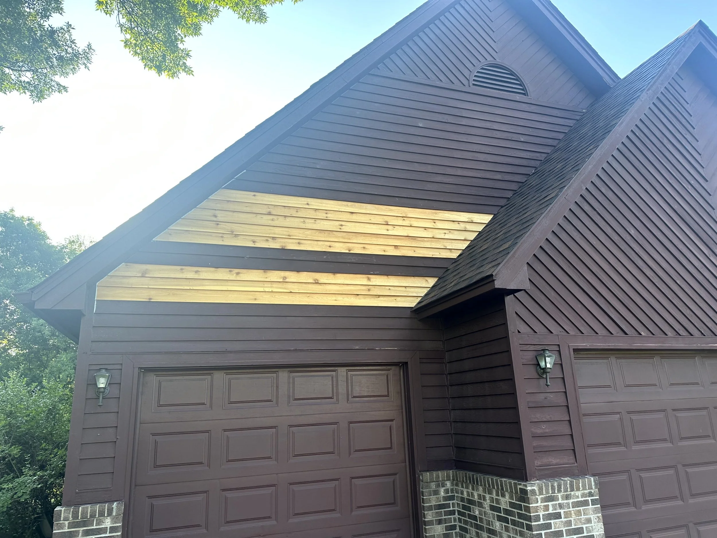 Close-up of a brown garage with a double door, exterior lights, a section of beige wood siding, and decorative brickwork at the base, under a clear blue sky with surrounding greenery.