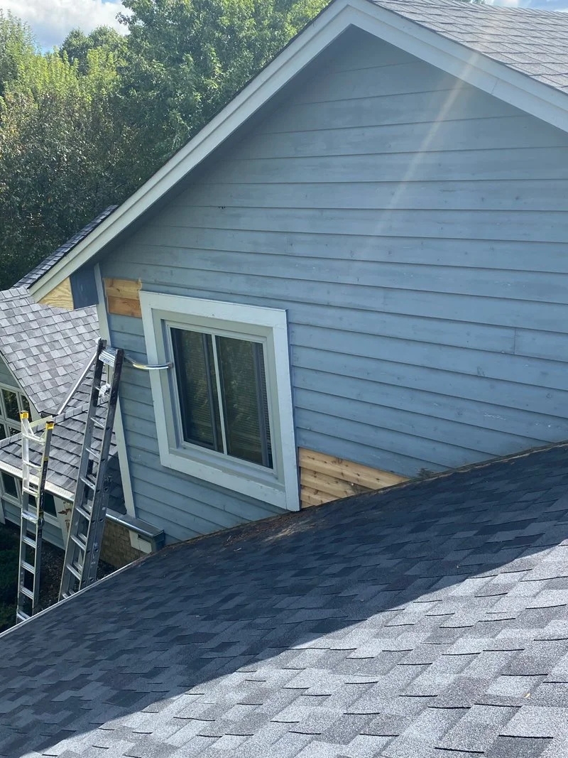View of a house under construction, showing light blue siding, a window with white trim, and gray roofing shingles. Ladder leaning against the house, with trees in the background.