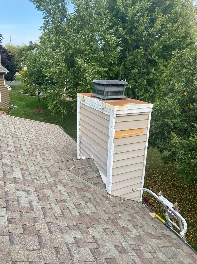 View of a chimney being repaired on a residential house roof with a vent cap, surrounded by green trees and neighboring houses, during daytime.
