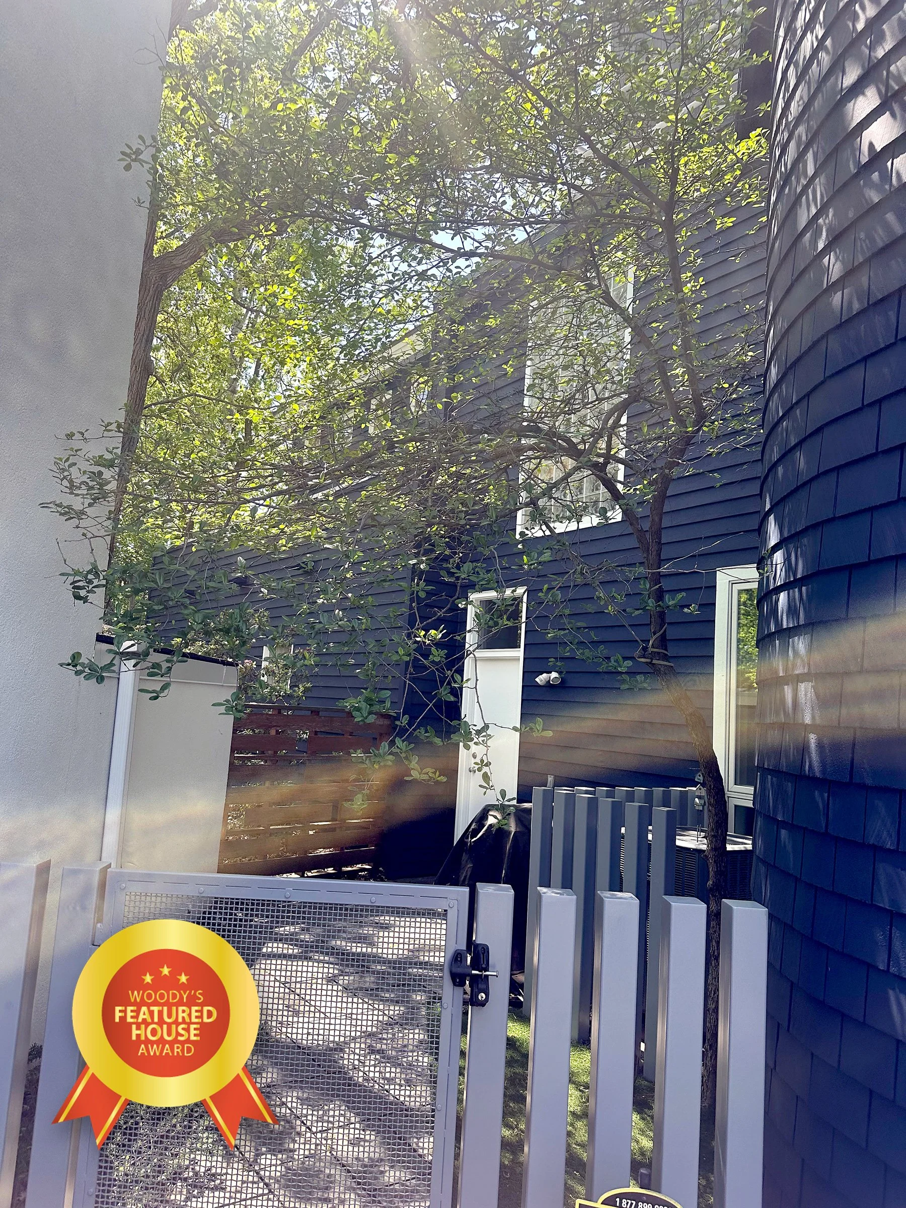 Exterior view of a modern house with dark blue siding, a tree growing close to the house, and a white gate with a WOODY'S FEATURED HOUSE AWARD sticker on it.